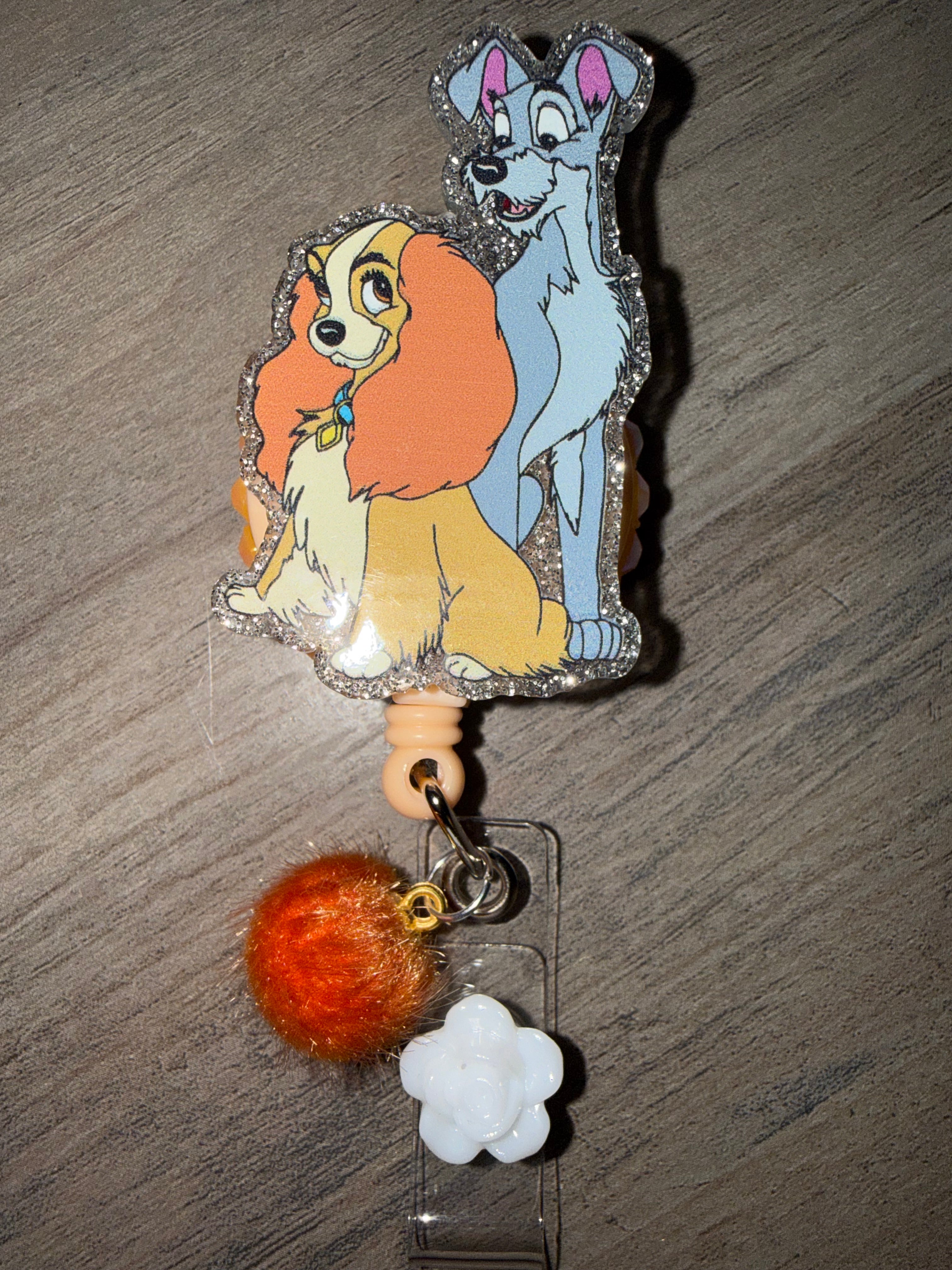 A glittery badge reel featuring Lady and the Tramp characters. The acrylic charm is bordered with silver glitter and attached to a tan retractable cord, decorated with an orange pom-pom and a small white flower charm hanging from the strap.