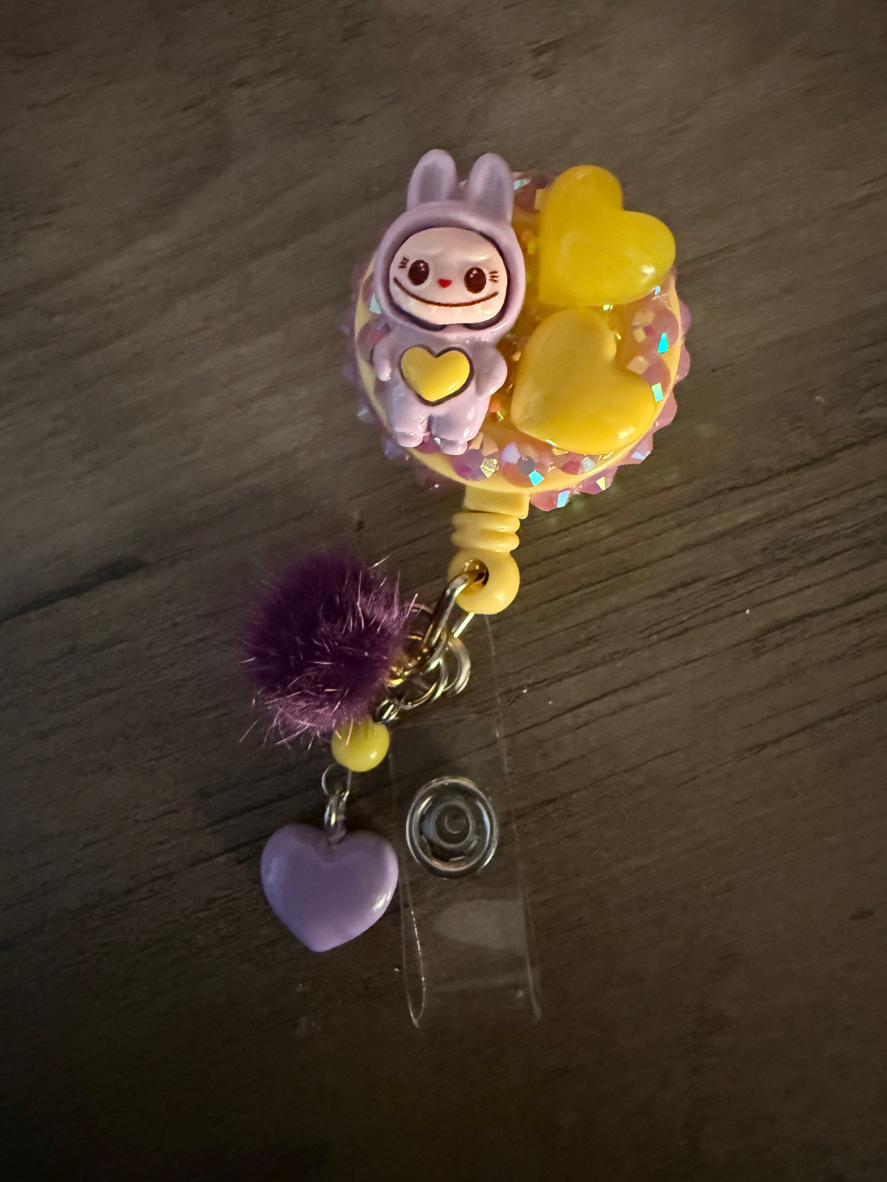 A purple and yellow handcrafted decoden badge reel featuring a small lavender monster figure in a hoodie, yellow resin hearts, and iridescent rhinestones, finished with a purple pom-pom and heart charm.