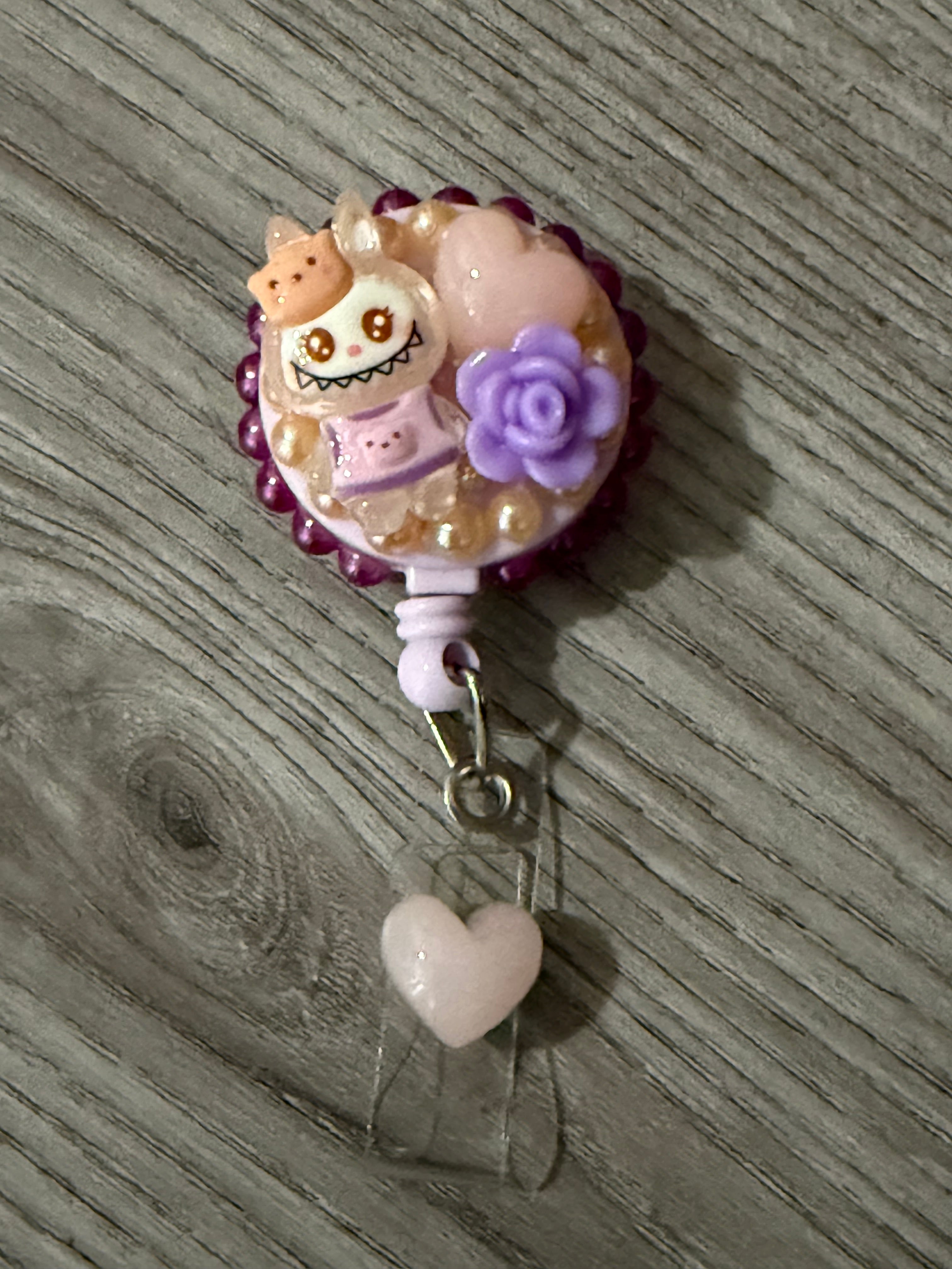 Cute badge reel featuring a whimsical monster with a crown, surrounded by pastel beads, a purple rose, and a pink heart charm.