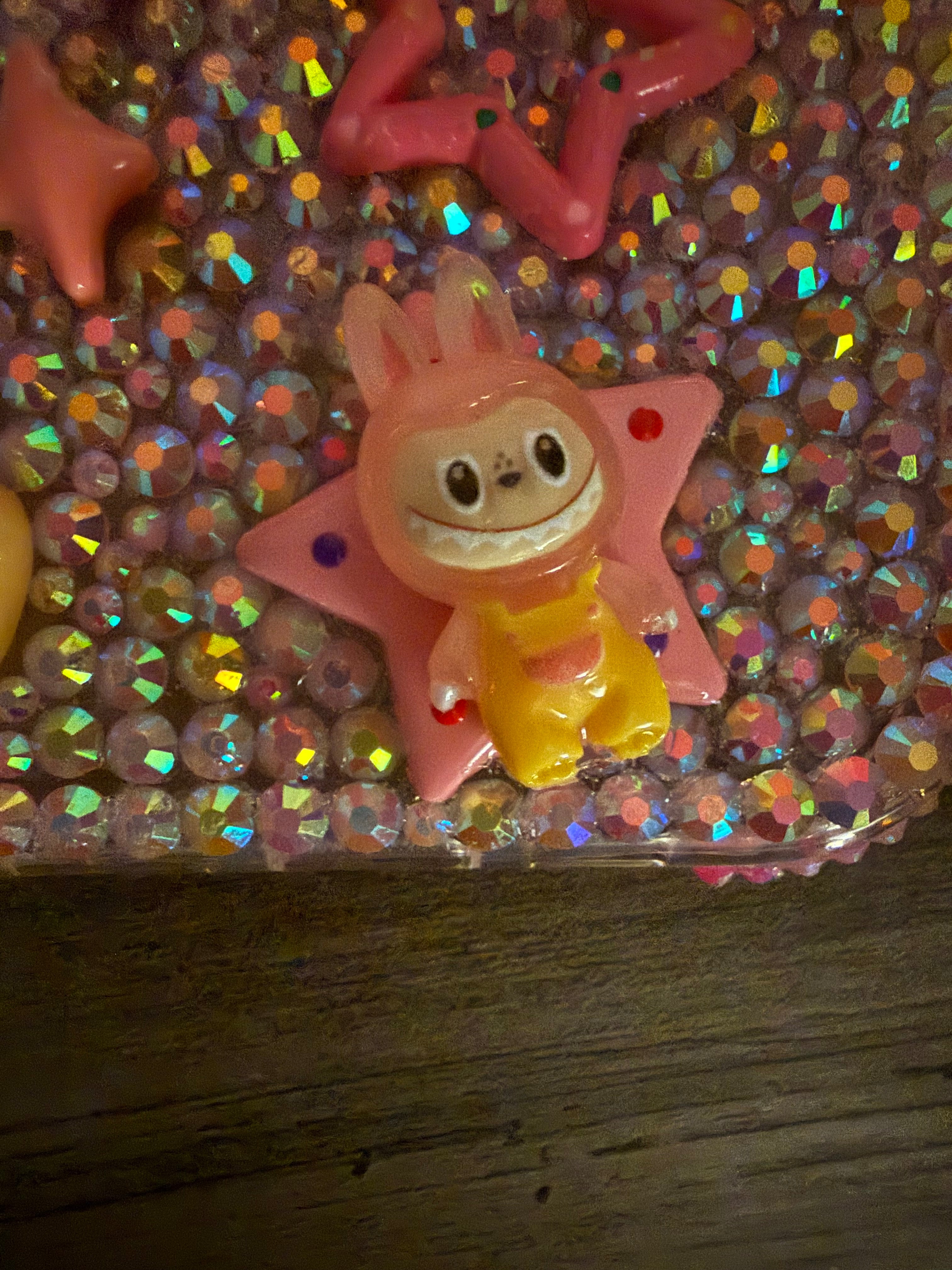 Close-up of a decoden phone case featuring iridescent rhinestones and a pink star cabochon with a cartoonish monster character charm wearing yellow overalls.