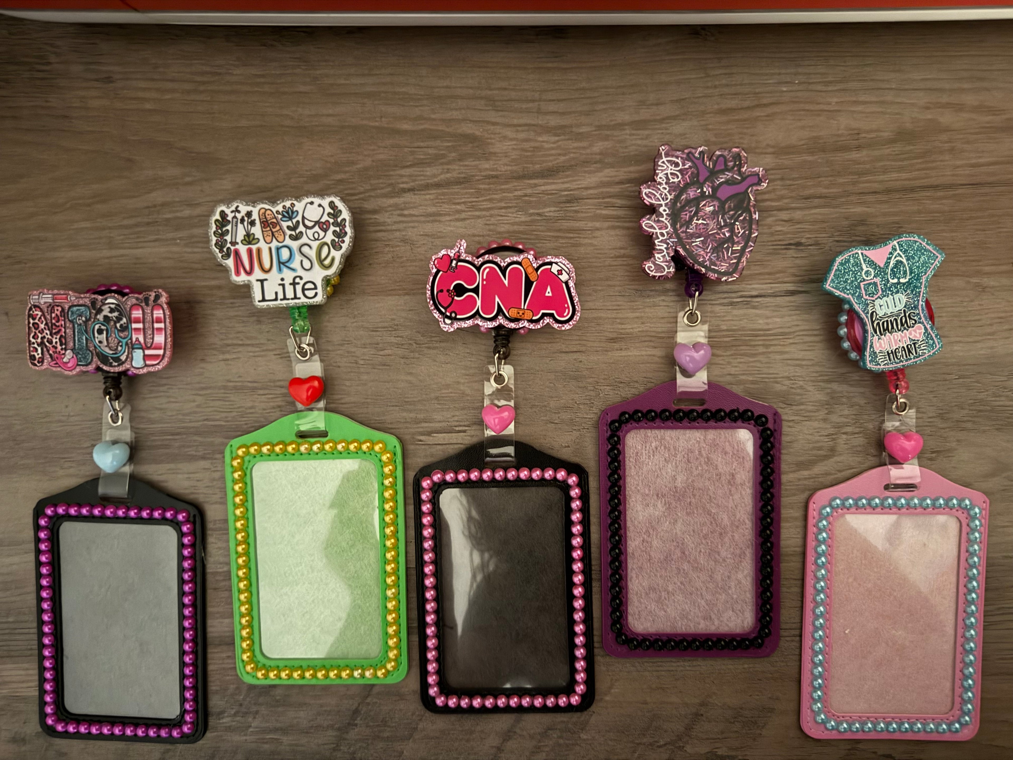 A row of five colorful, decorative badge reels and matching ID holders featuring medical themes and "decoden" style embellishments like glitter, small pearls, and heart charms. The designs include text for "NICU," "Nurse Life," "CNA," a sparkling anatomical heart for "Cardiology," and a scrub top graphic that reads "Cold Hands Warm Heart."