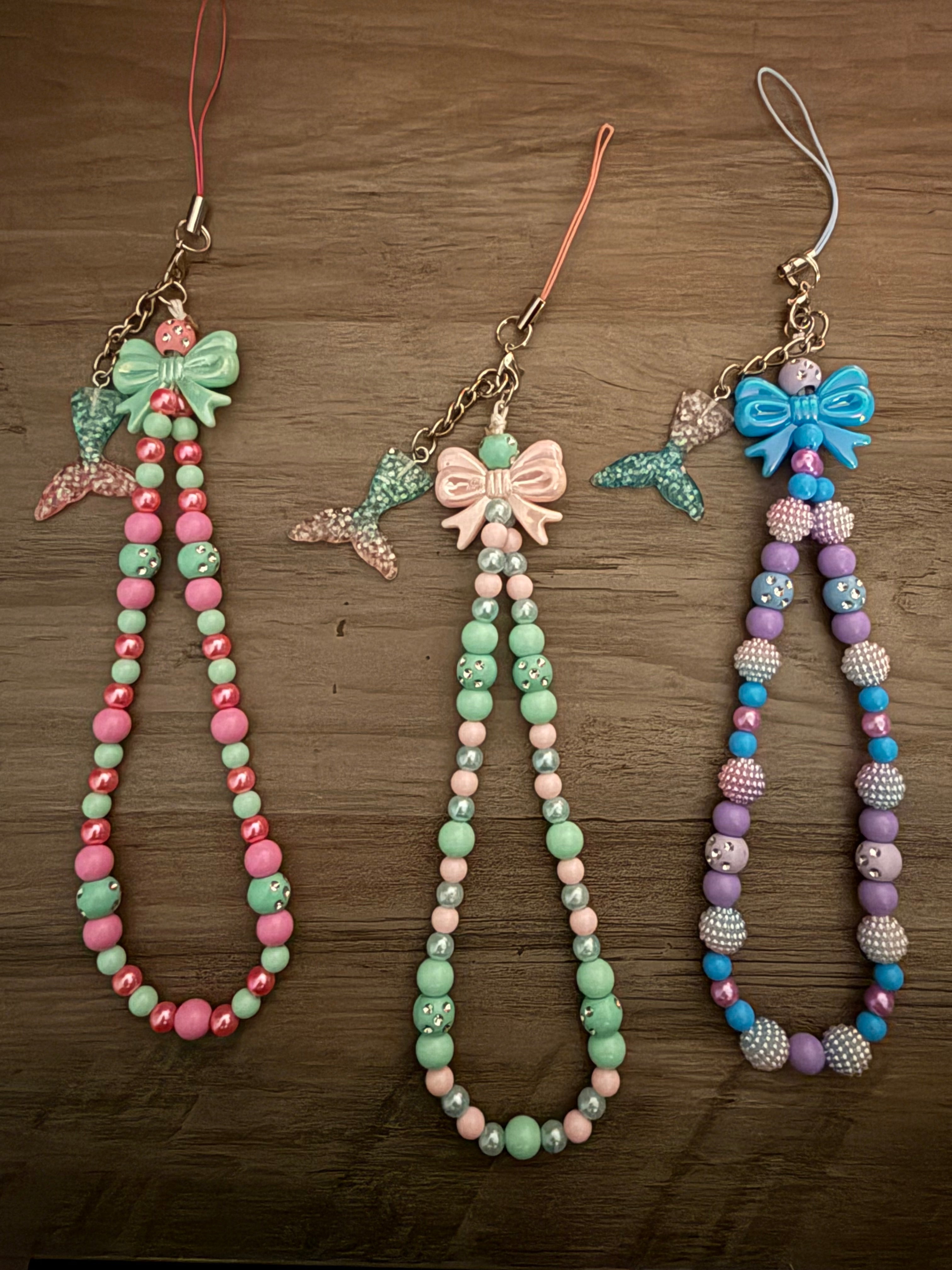 Three beaded phone wristlet straps in pastel pink/green, mint/peach, and blue/purple, each decorated with a large bow and a dangling mermaid tail charm.
