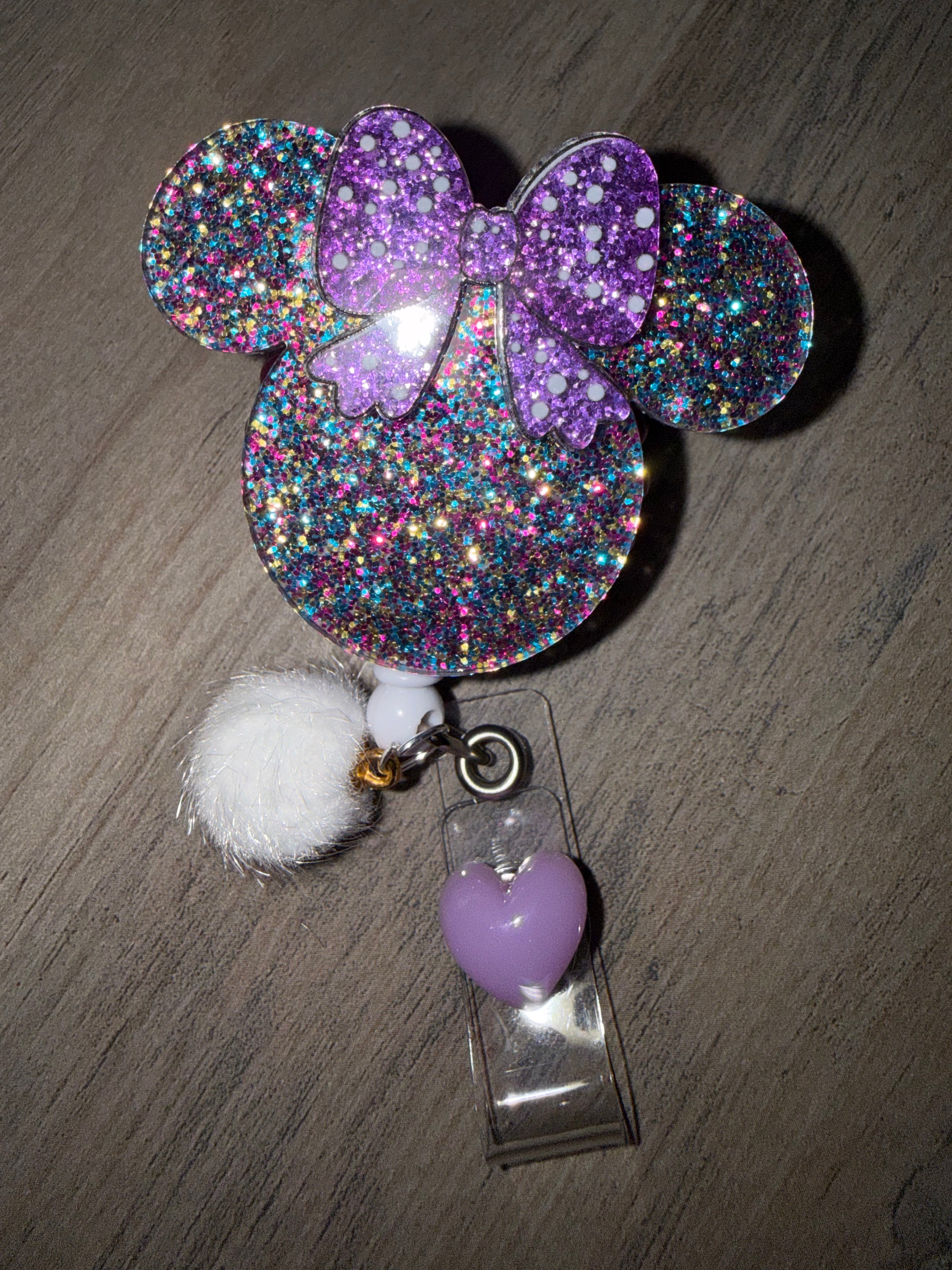 A glittery badge reel in the shape of a Minnie Mouse head, featuring multicolored chunky glitter and a purple polka-dot bow. The retractable clip is decorated with a white pom-pom and a small lilac heart charm hanging from the clear strap.