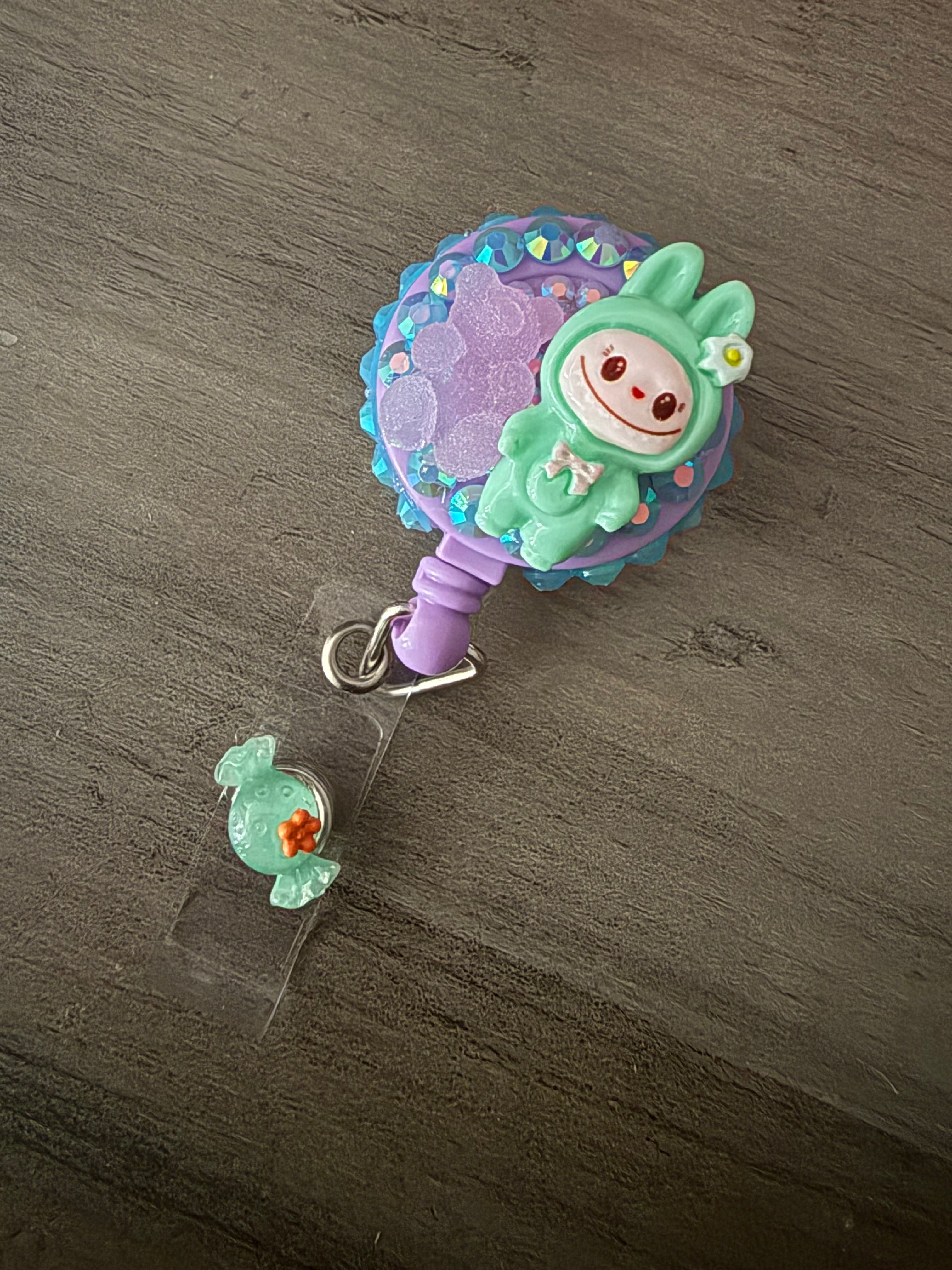 Handcrafted mint green monster in a hooded outfit on a purple retractable badge reel, embellished with blue and iridescent rhinestones, a purple gummy bear charm, and a mint candy charm on the strap.