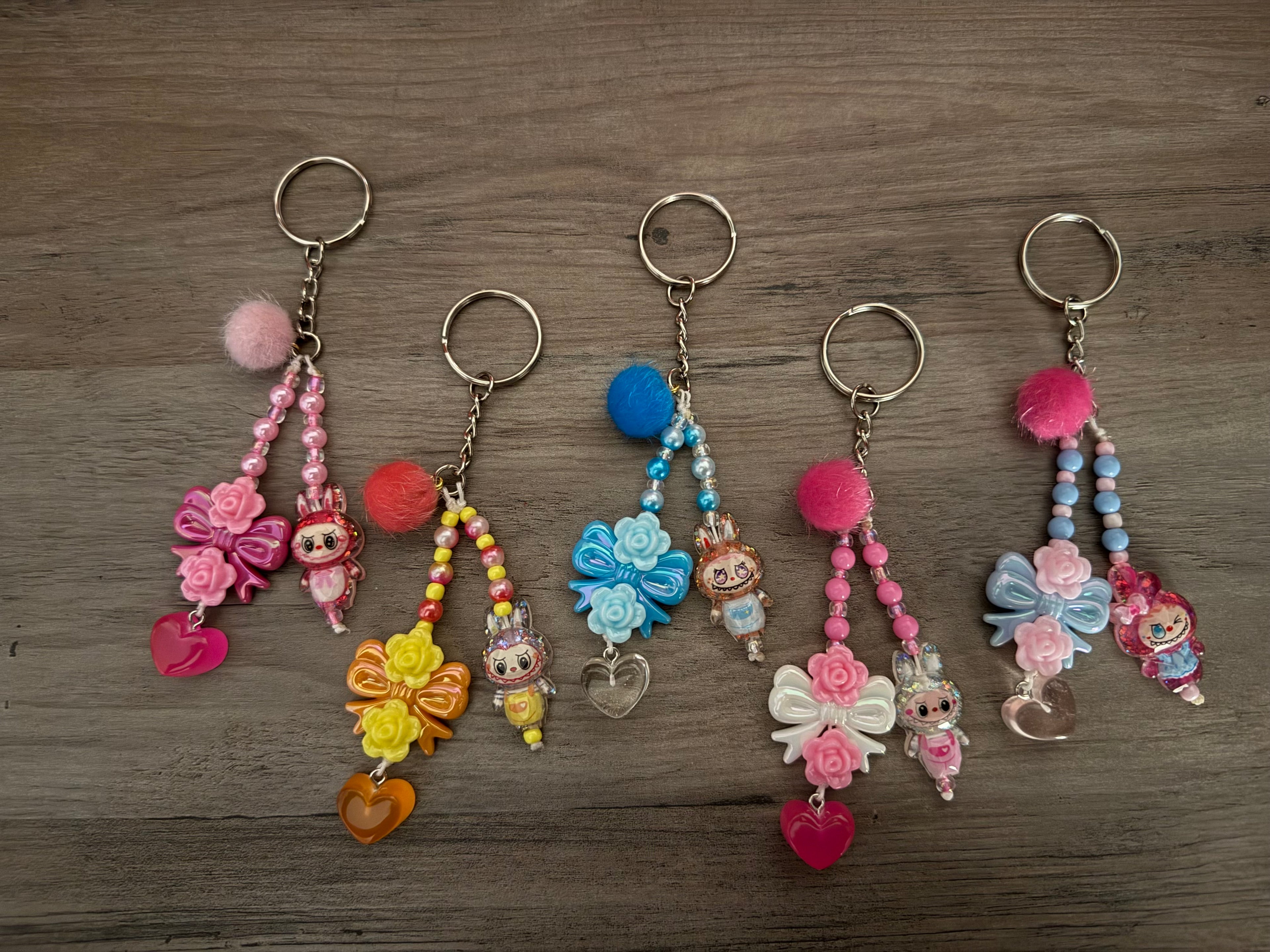 A row of five handcrafted "kawaii" style keychains on a wooden surface. Each keychain features a combination of colorful beads, pom-poms, floral bows, heart charms, and small monster figurines in various pastel shades of pink, yellow, blue, and white.