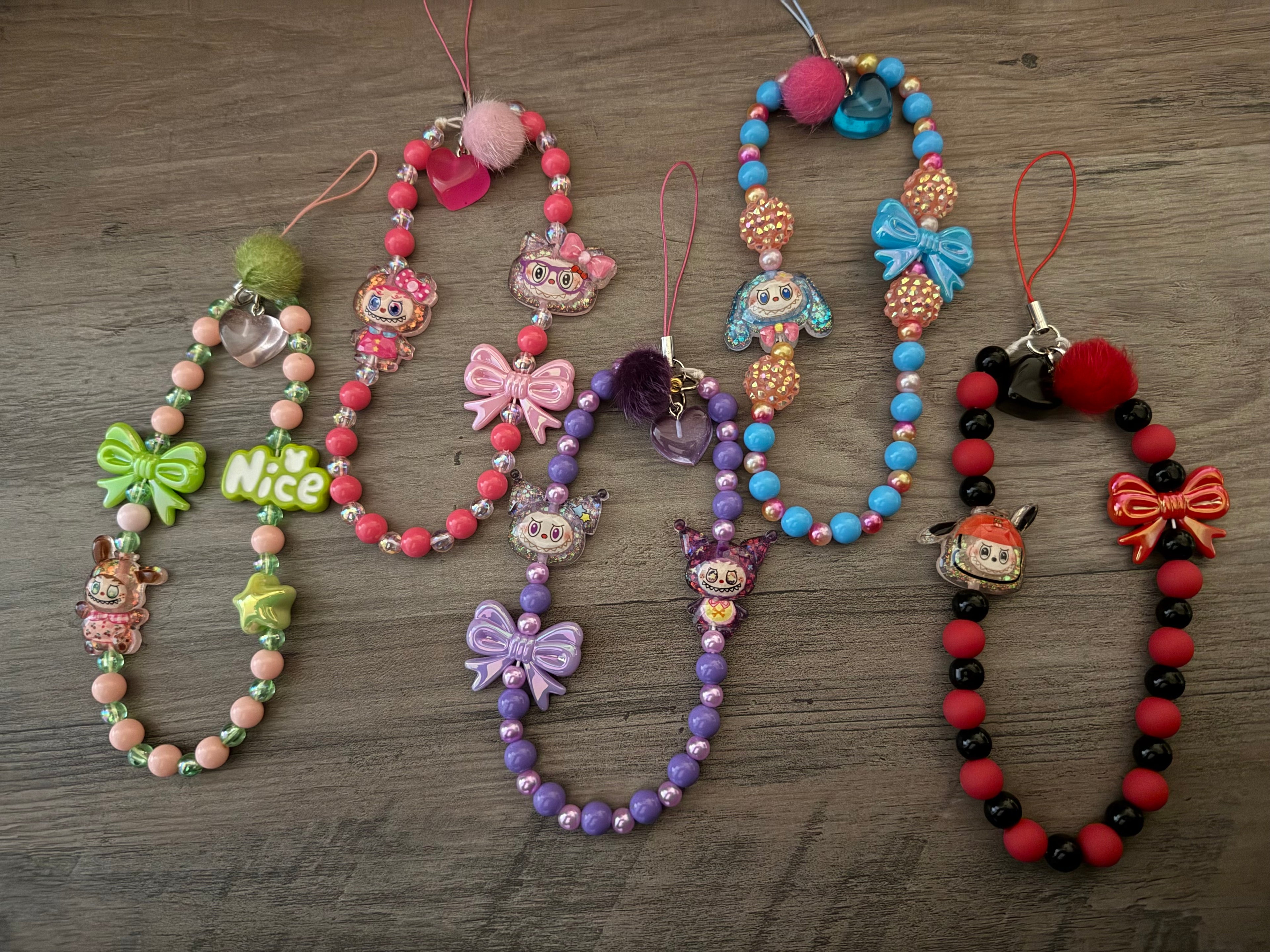 A collection of five colorful, beaded phone charms featuring pom-poms, bows, and character charms in green, pink, purple, blue, and red themes.