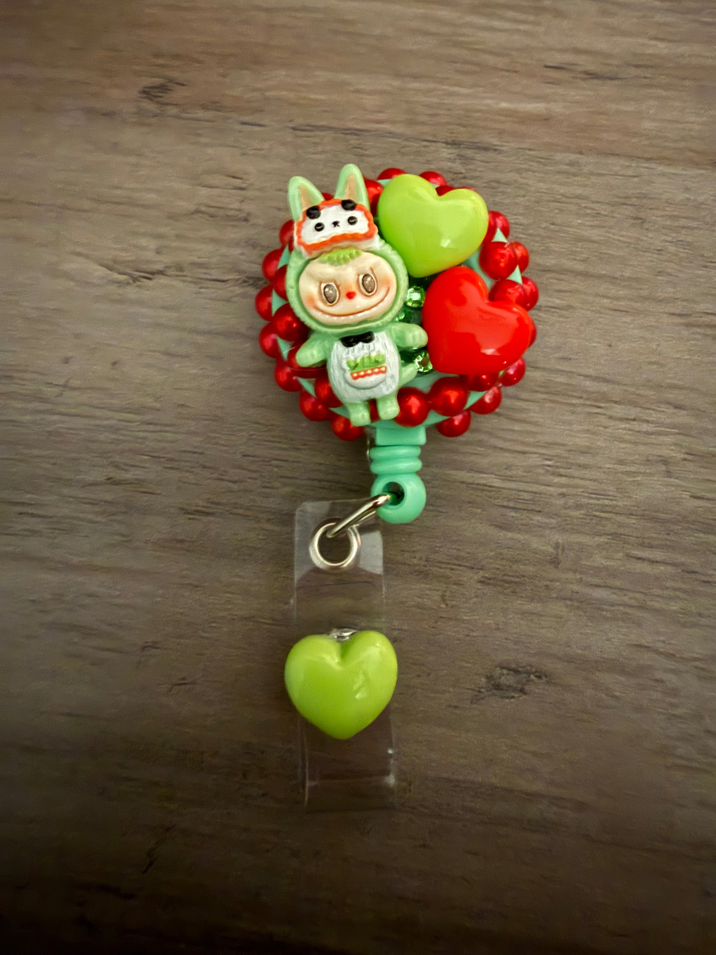 A decorative mint green badge reel featuring a monster figure in a green bunny outfit, embellished with red pearls and lime green and red hearts.