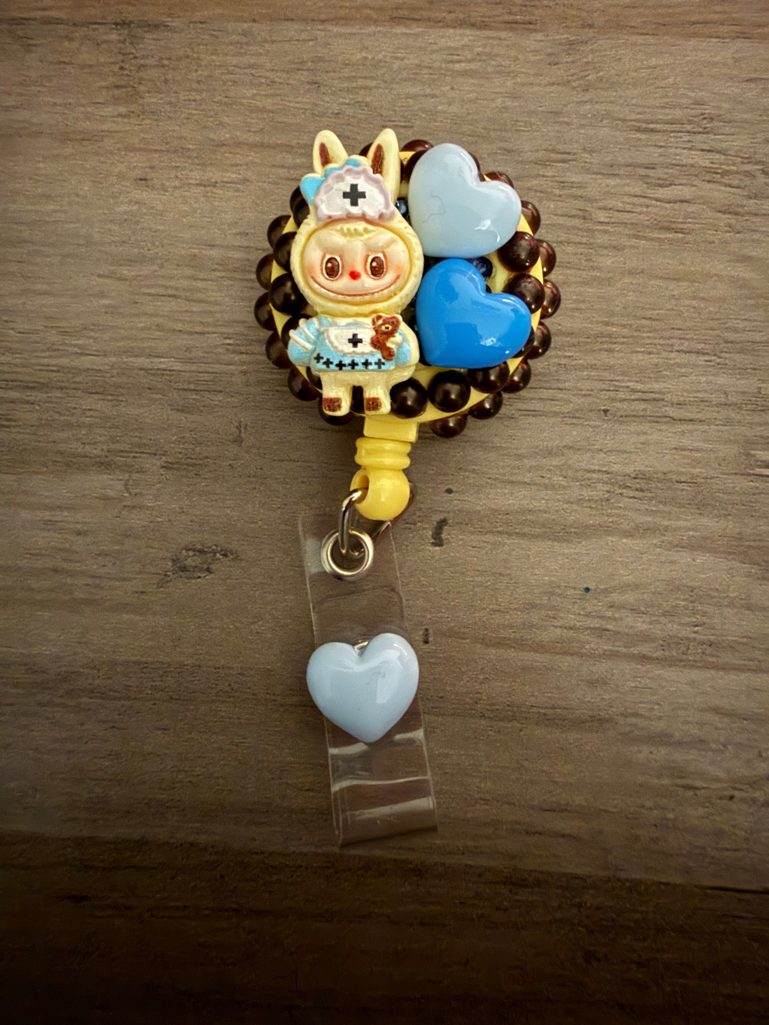 A decorative yellow badge reel featuring a monster figure in a nurse-themed bunny outfit, embellished with dark brown pearls and light and dark blue hearts.