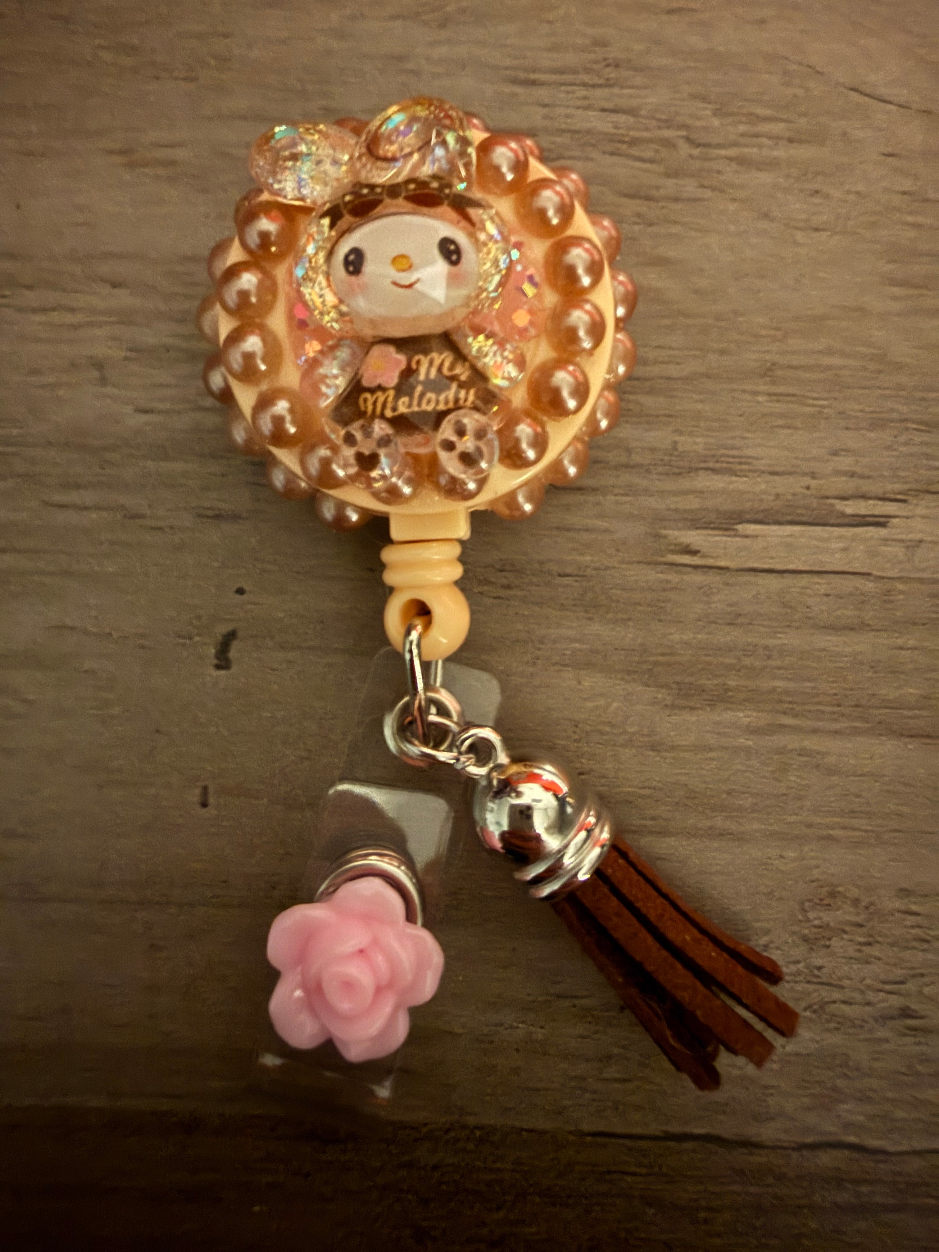 A decoden retractable badge reel featuring a My Melody character surrounded by bronze-toned pearls, accented with a brown suede tassel and a pink rose charm.