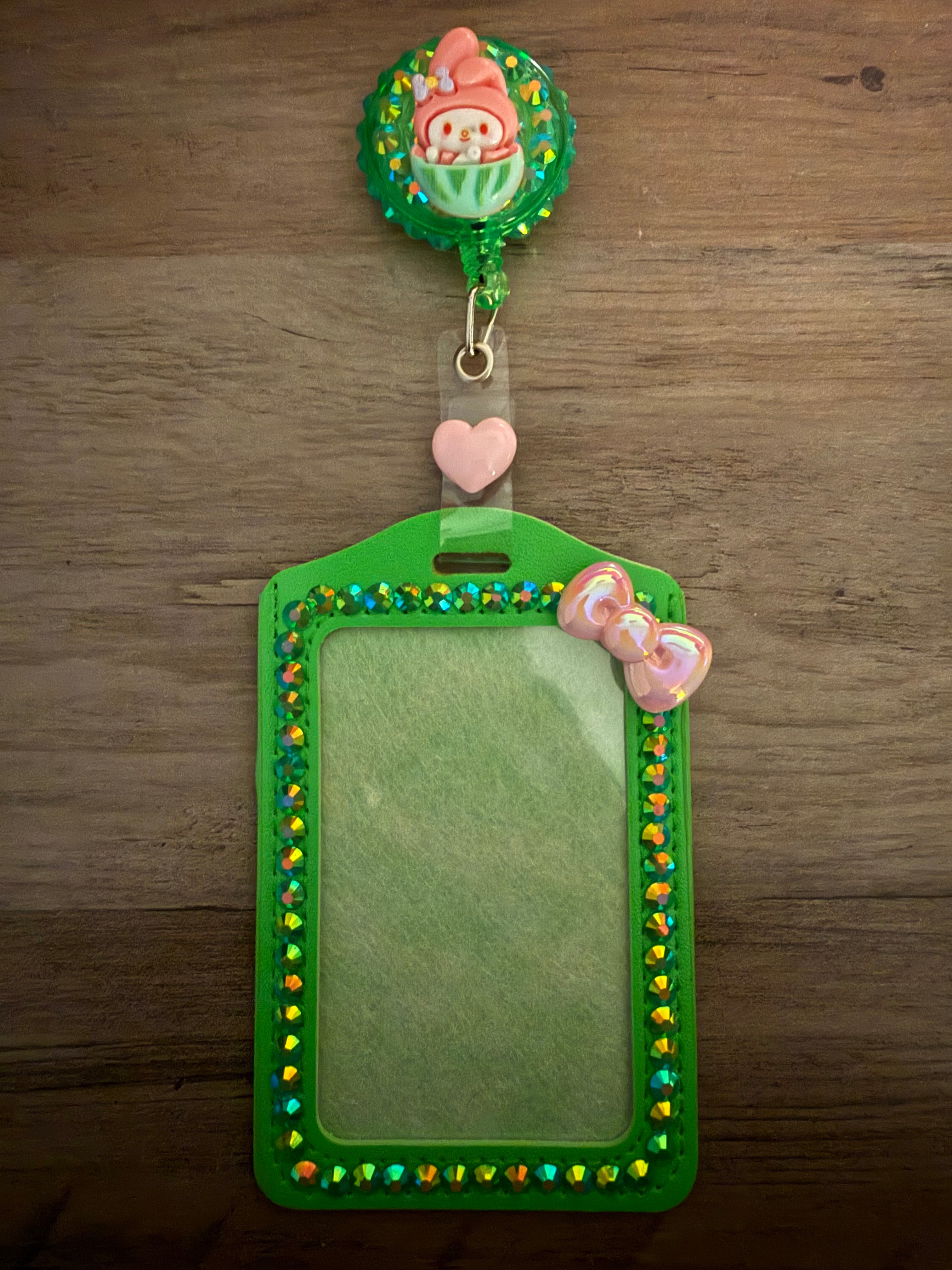 A green, rhinestone-embellished ID card holder with a matching retractable badge reel. The reel features a My Melody character charm sitting in a green cup against a glittering background, and the holder is lined with iridescent green gems and a small pink pearlescent bow.