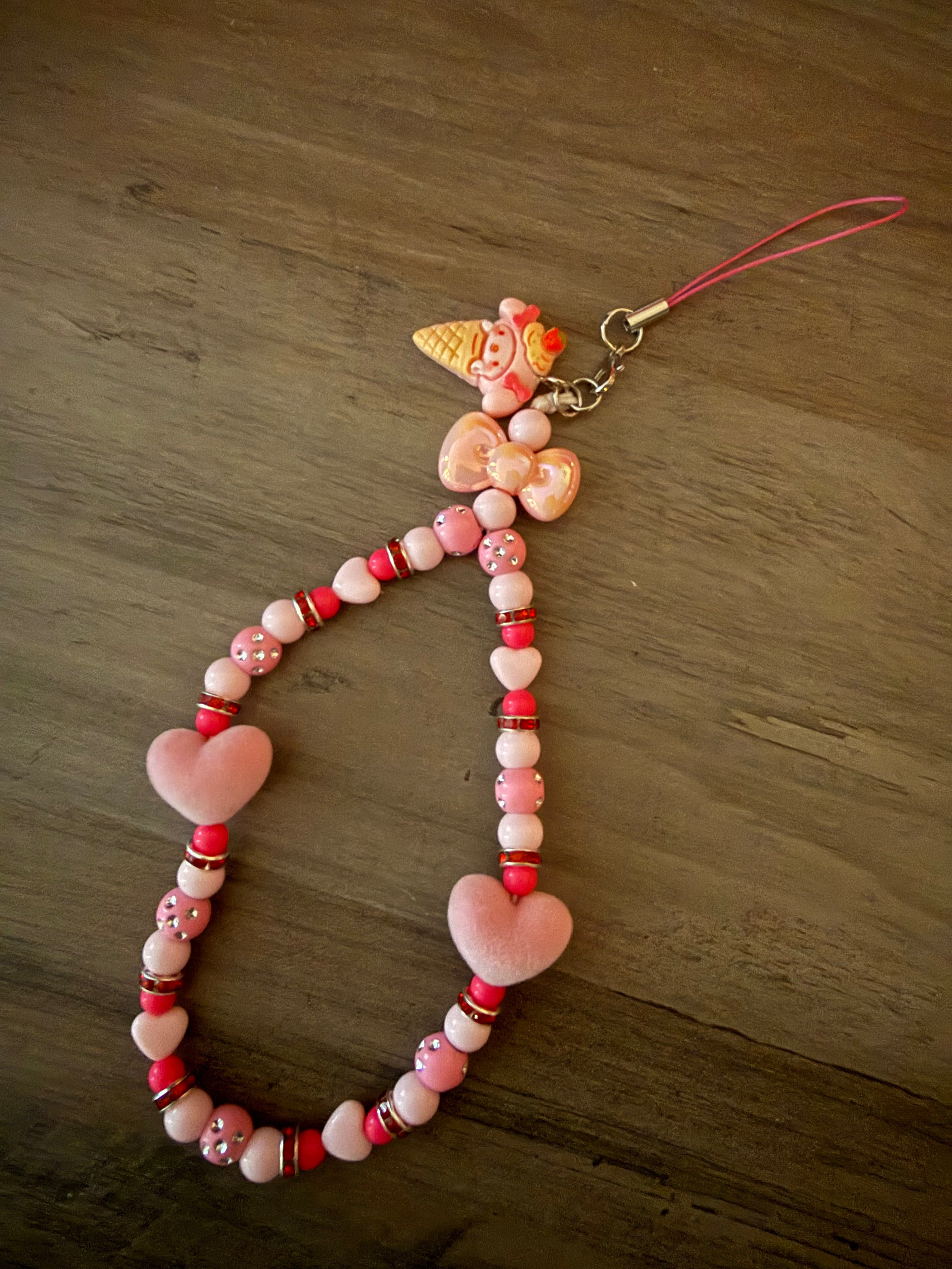 A handmade beaded phone wristlet featuring various shades of pink and red beads, sparkling rhinestone spacers, pink heart charms, a peach bow, and a My Melody ice cream cone charm.