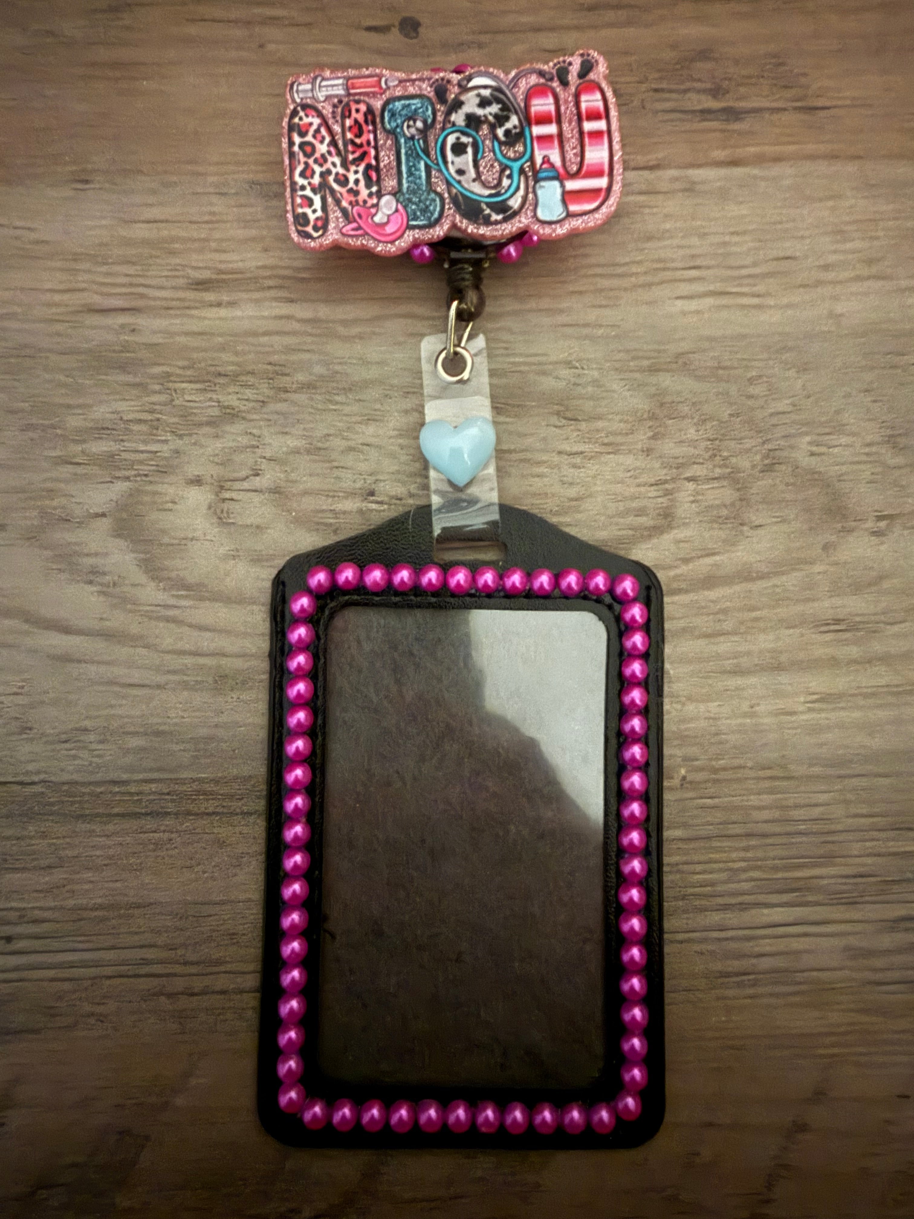 A handmade NICU-themed badge reel featuring pink glitter and patterns like leopard print and stripes. The reel is attached to a black ID holder lined with small, hot pink decorative pearls and accented with a light blue heart charm.