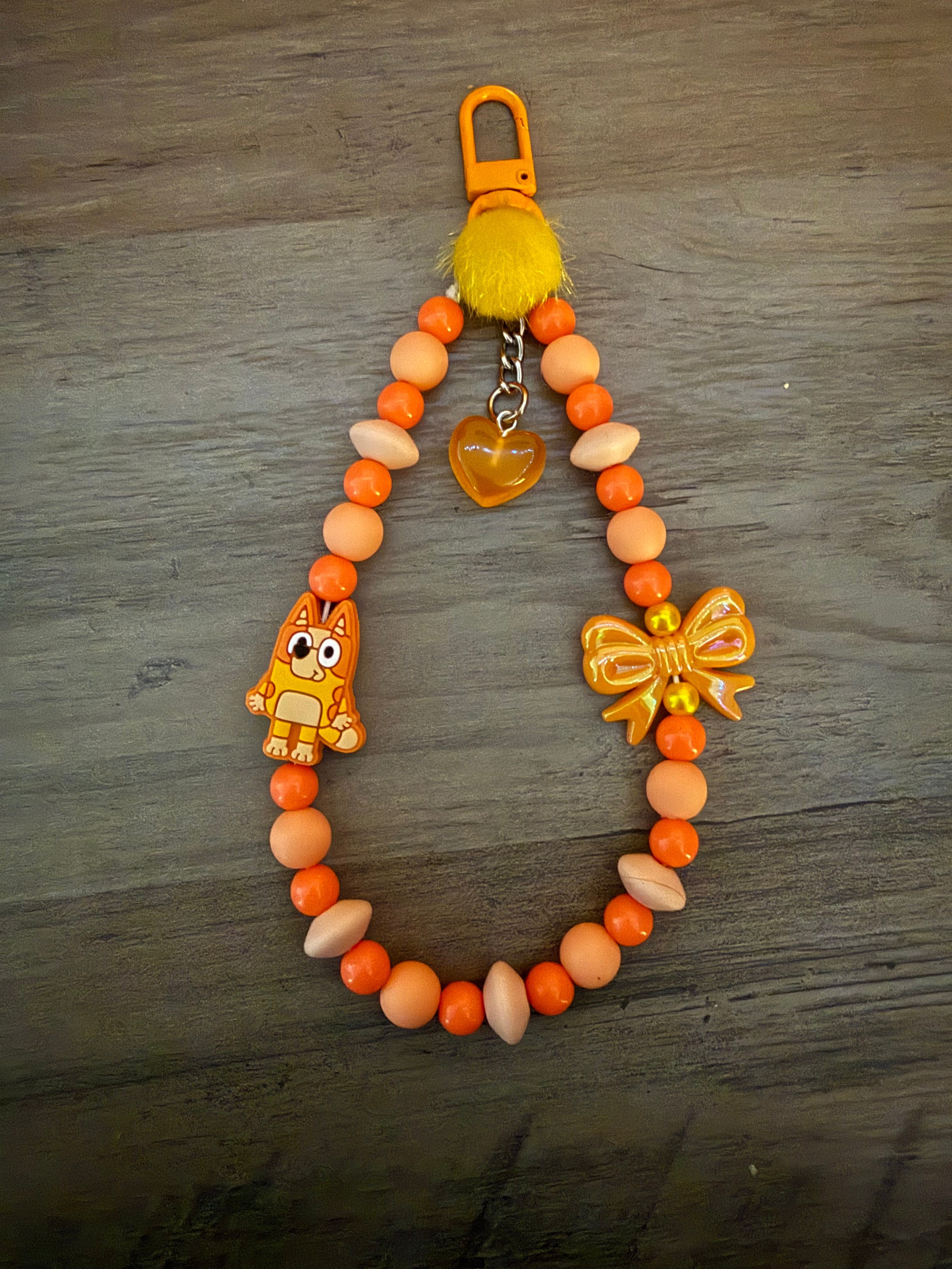 An orange beaded lanyard featuring shades of orange and peach beads, a yellow pom-pom, a metallic orange bow, an orange heart charm, and a small character charm of an orange cartoon dog.