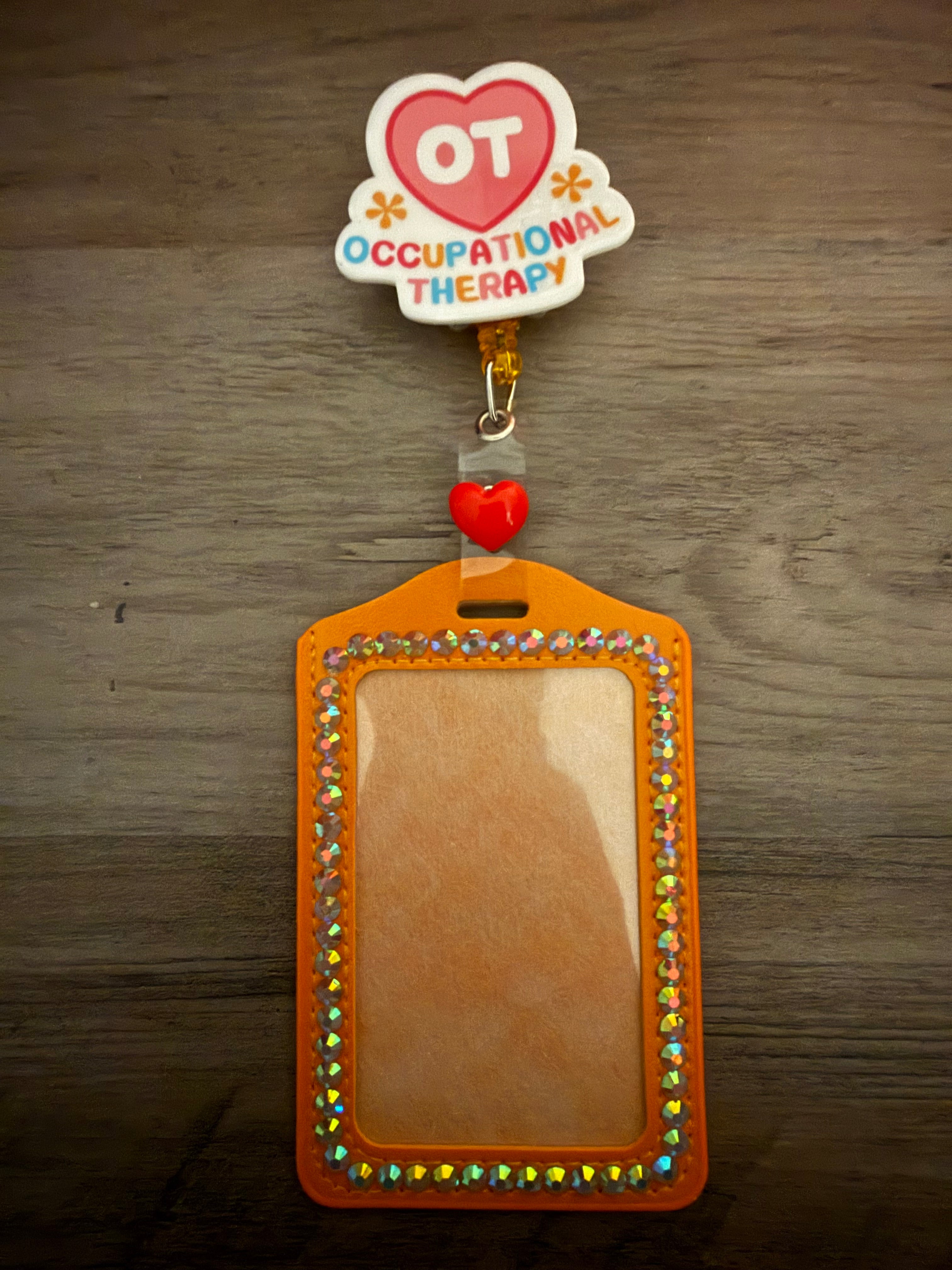 An orange ID badge holder lined with iridescent rhinestones, connected to a retractable reel featuring a pink heart with the letters "OT" and the text "OCCUPATIONAL THERAPY" in colorful letters.