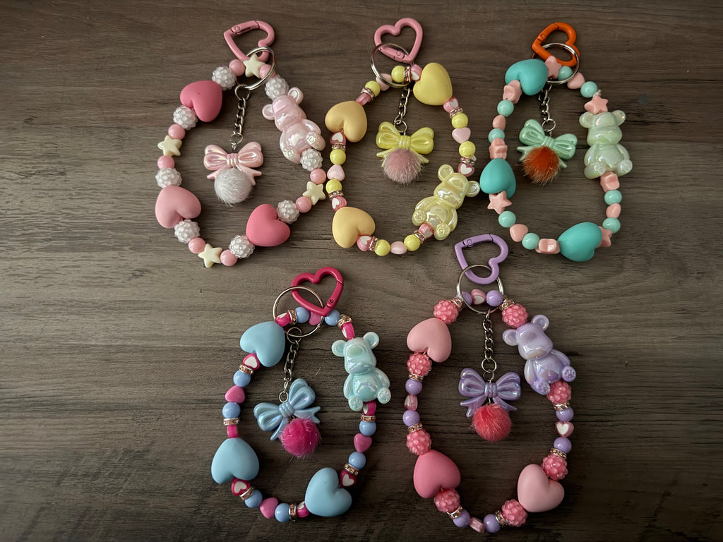 Five handcrafted pastel beaded wristlet keychains in shades of pink, yellow, teal, blue, and lavender. Each keychain features large heart-shaped beads, a iridescent gummy bear charm, and a central bow charm with a fuzzy pom-pom, all attached to a heart-shaped clasp.