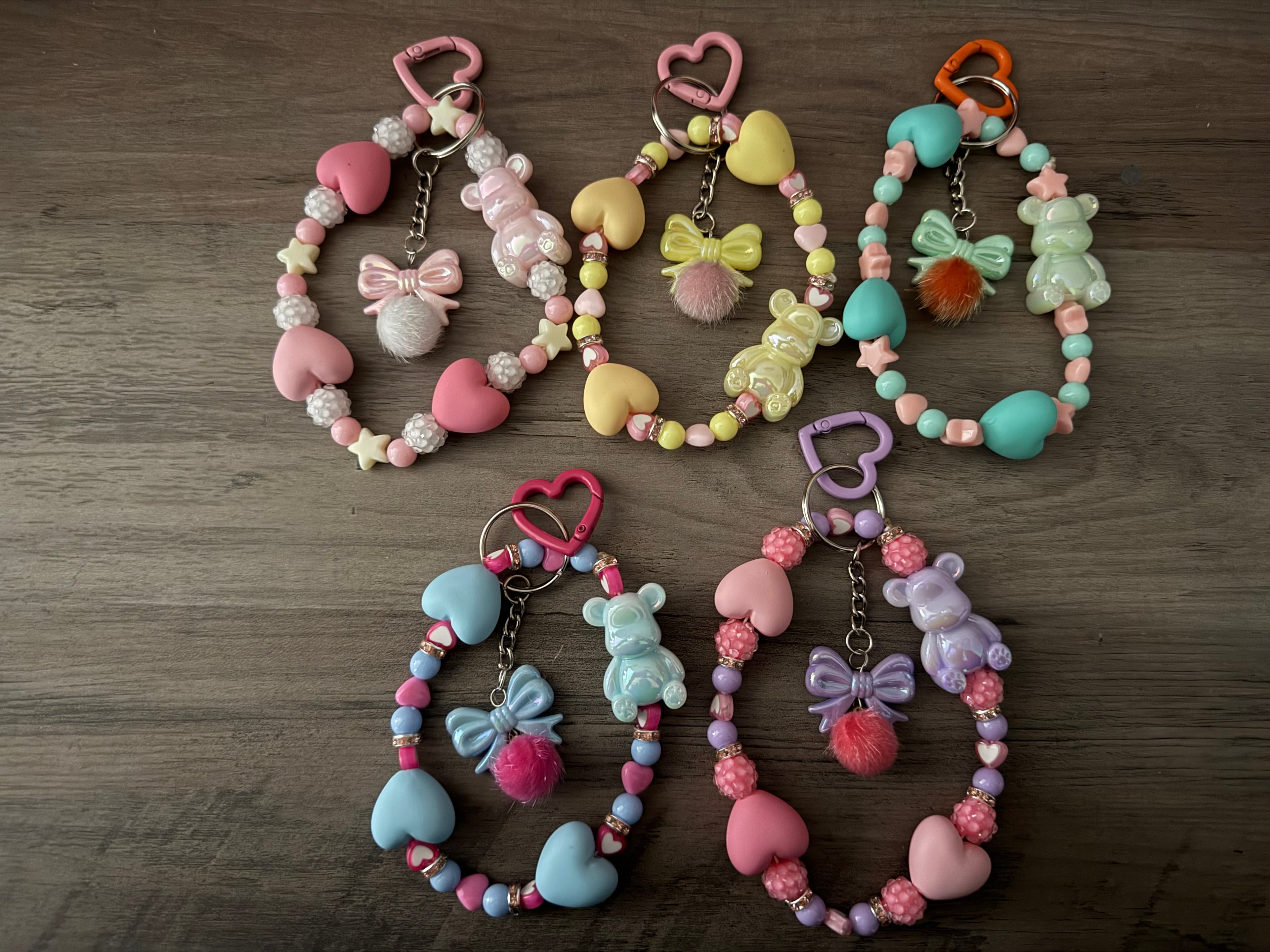 Five handcrafted pastel beaded wristlet keychains in shades of pink, yellow, teal, blue, and lavender. Each keychain features large heart-shaped beads, a iridescent gummy bear charm, and a central bow charm with a fuzzy pom-pom, all attached to a heart-shaped clasp.