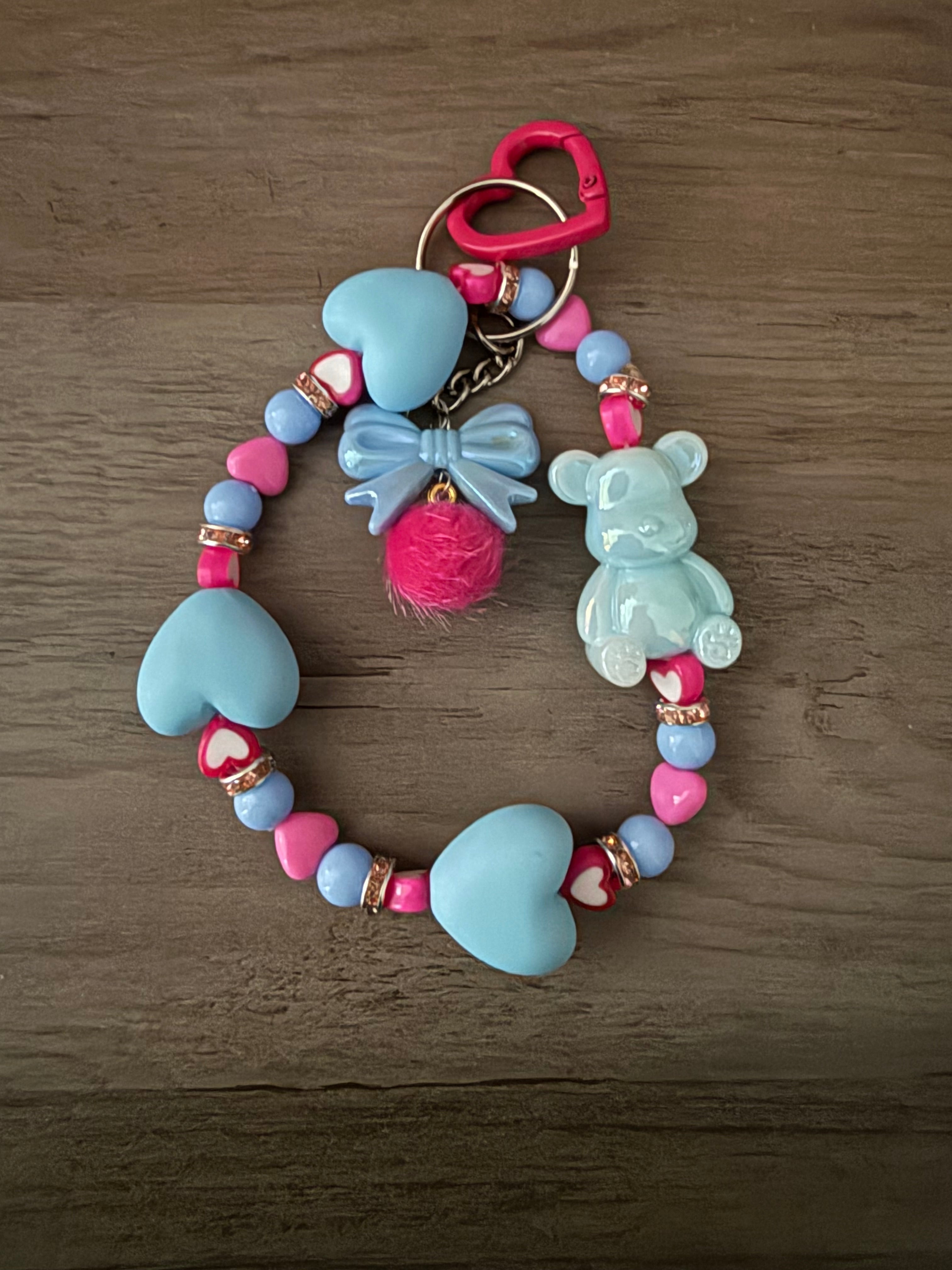 A handcrafted blue and hot pink beaded wristlet keychain featuring matte light blue heart-shaped beads, an iridescent blue gummy bear charm, and a central blue bow with a bright pink fuzzy pom-pom. The design includes small pink heart beads and gold-toned rhinestone spacers, all attached to a hot pink heart-shaped clasp.