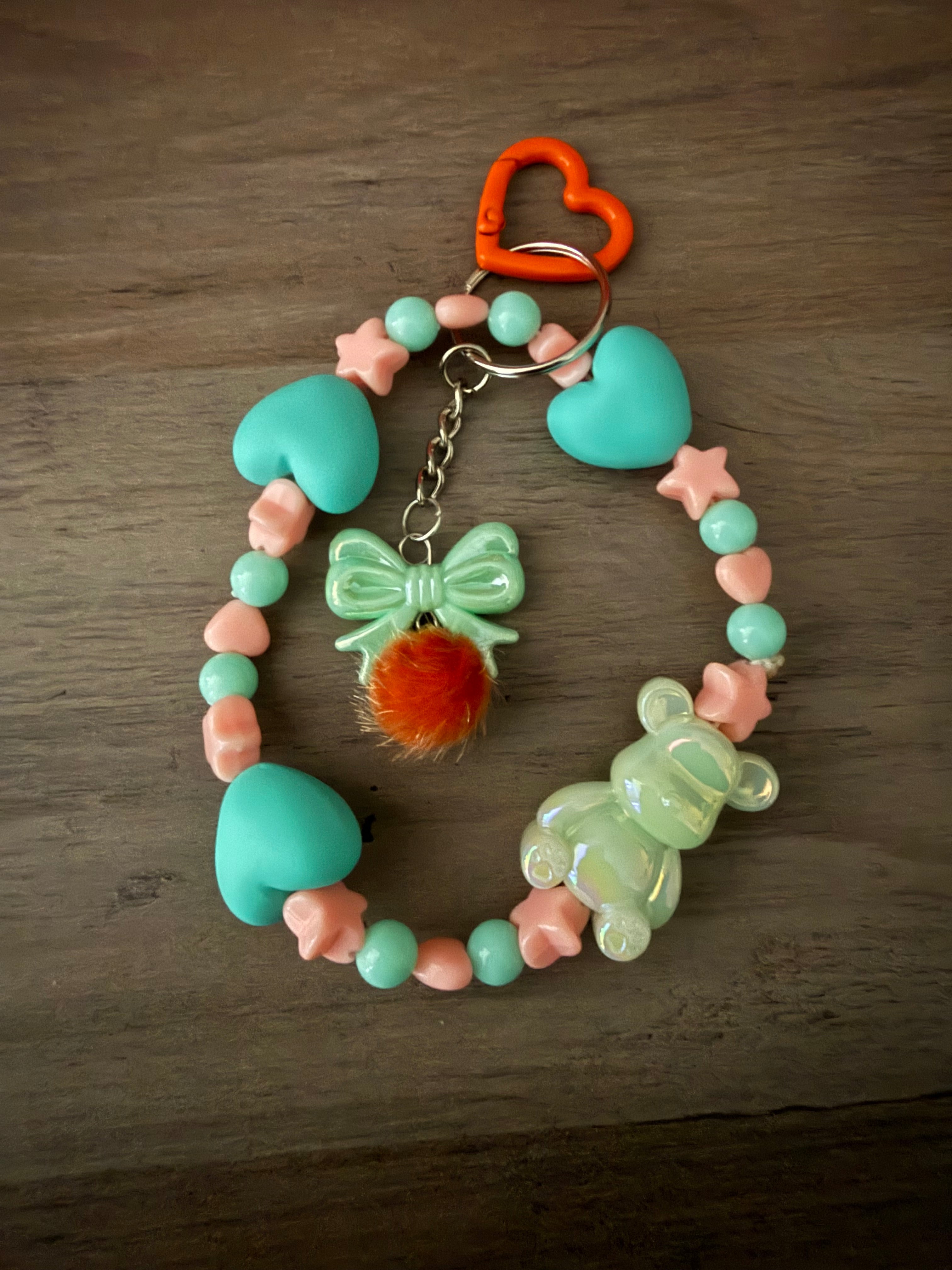 A handcrafted teal and peach beaded wristlet keychain featuring matte teal heart-shaped beads, peach star beads, and an iridescent mint green gummy bear charm. The center features a mint green bow with an orange fuzzy pom-pom, all hanging from an orange heart-shaped clasp.