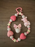 A handcrafted pink beaded wristlet keychain featuring matte heart-shaped beads, iridescent gummy bear and star charms, and a center pink bow with a white fuzzy pom-pom. The beads are a mix of smooth pink and textured white, all attached to a heart-shaped pink clasp.
