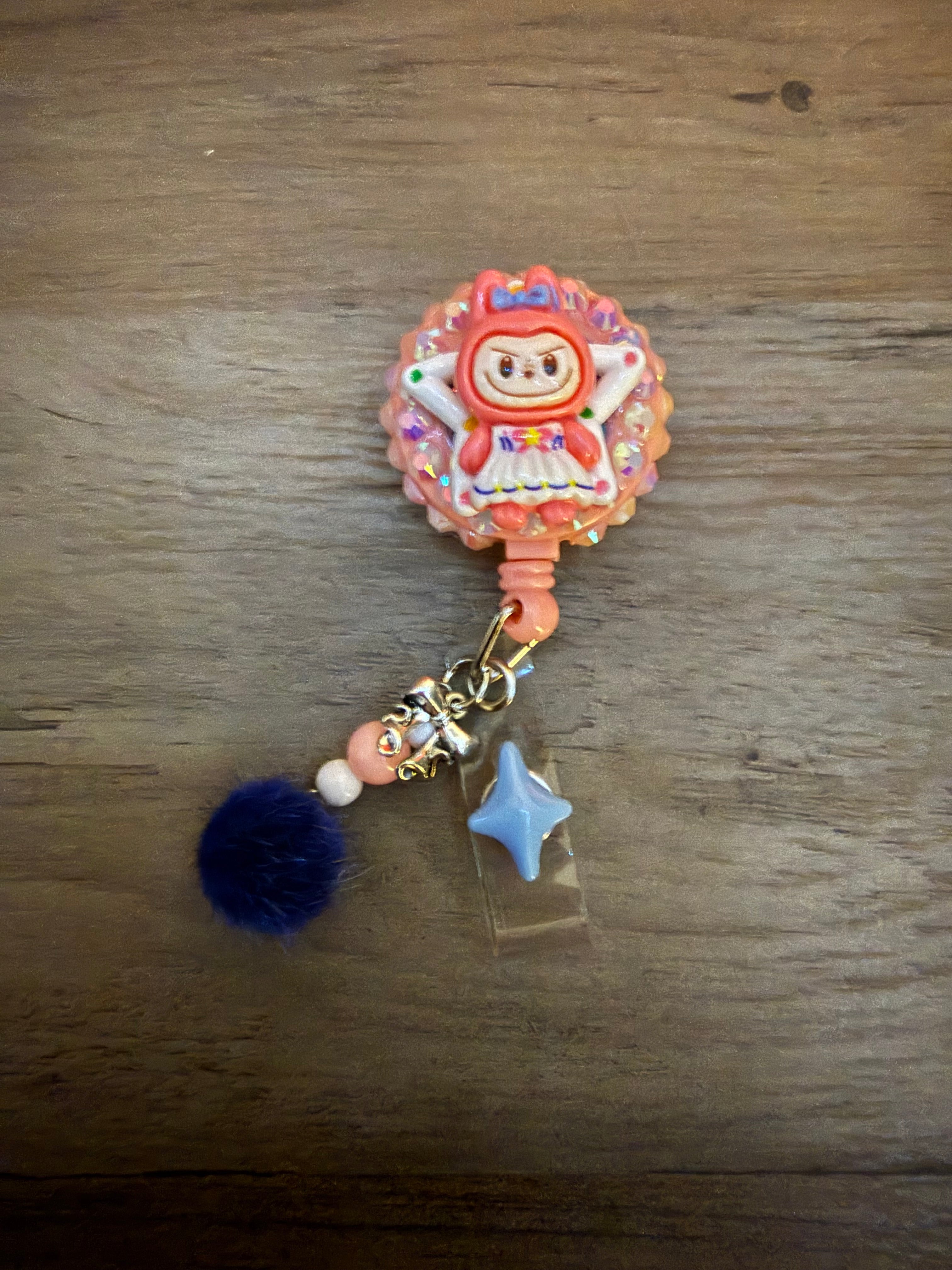 A single, detailed handmade badge reel featuring an anime-style peach monster with blue eyes and a playful pose. The character is set on a star-shaped base surrounded by sparkling gems. The reel includes dangling accessories such as pink and pearl beads, a silver bow charm, a light blue star, and a small, fuzzy dark blue pom-pom on a dark wood grain background.