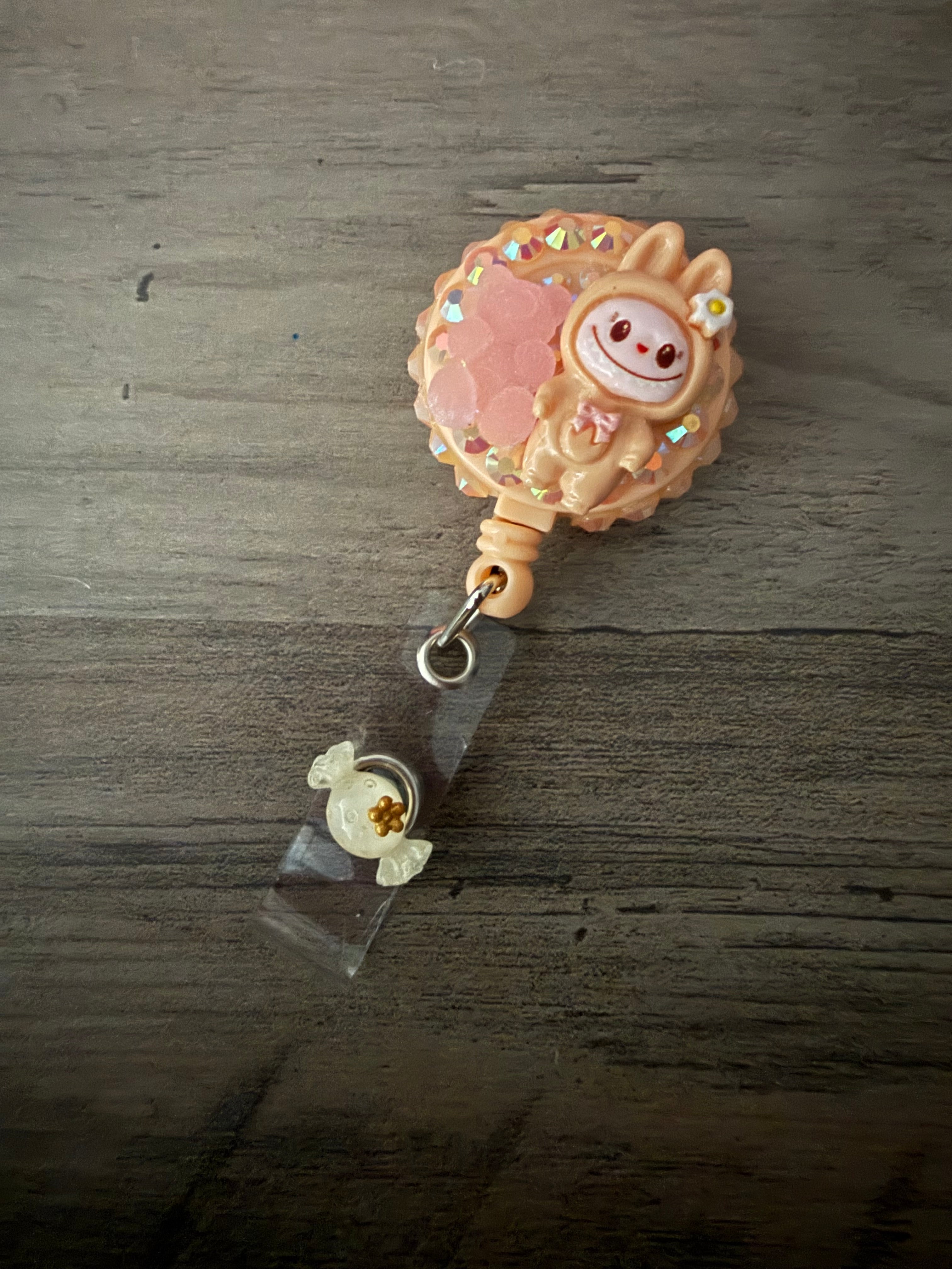 Handcrafted peach-colored monster in a hooded outfit on a matching retractable badge reel, featuring iridescent rhinestones, a pink gummy bear charm, and a translucent cream candy charm on the strap.