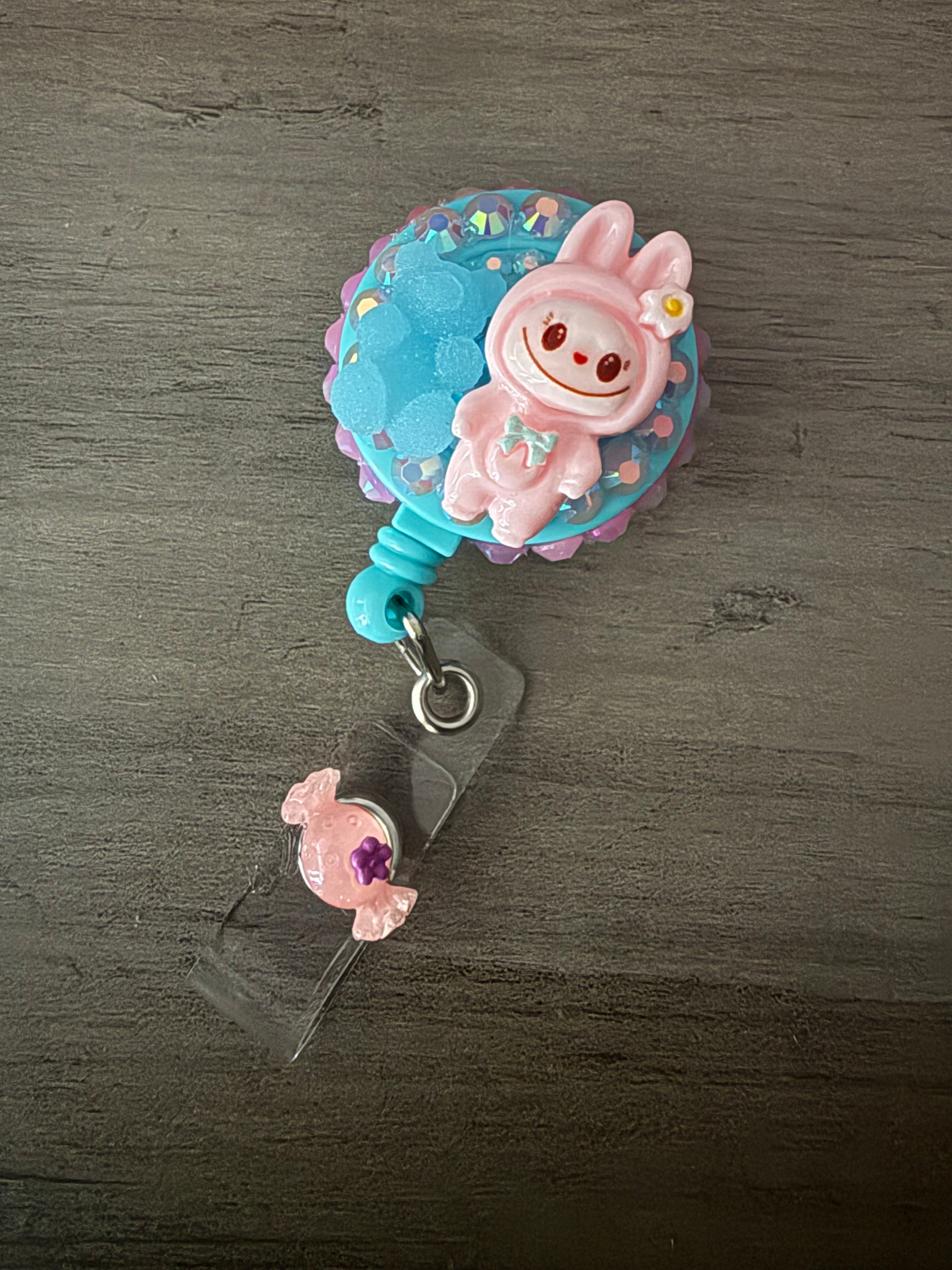 Handcrafted pink monster in a hooded outfit on a light blue retractable badge reel, accented with iridescent rhinestones, a blue gummy bear charm, and a small pink candy charm on the strap.