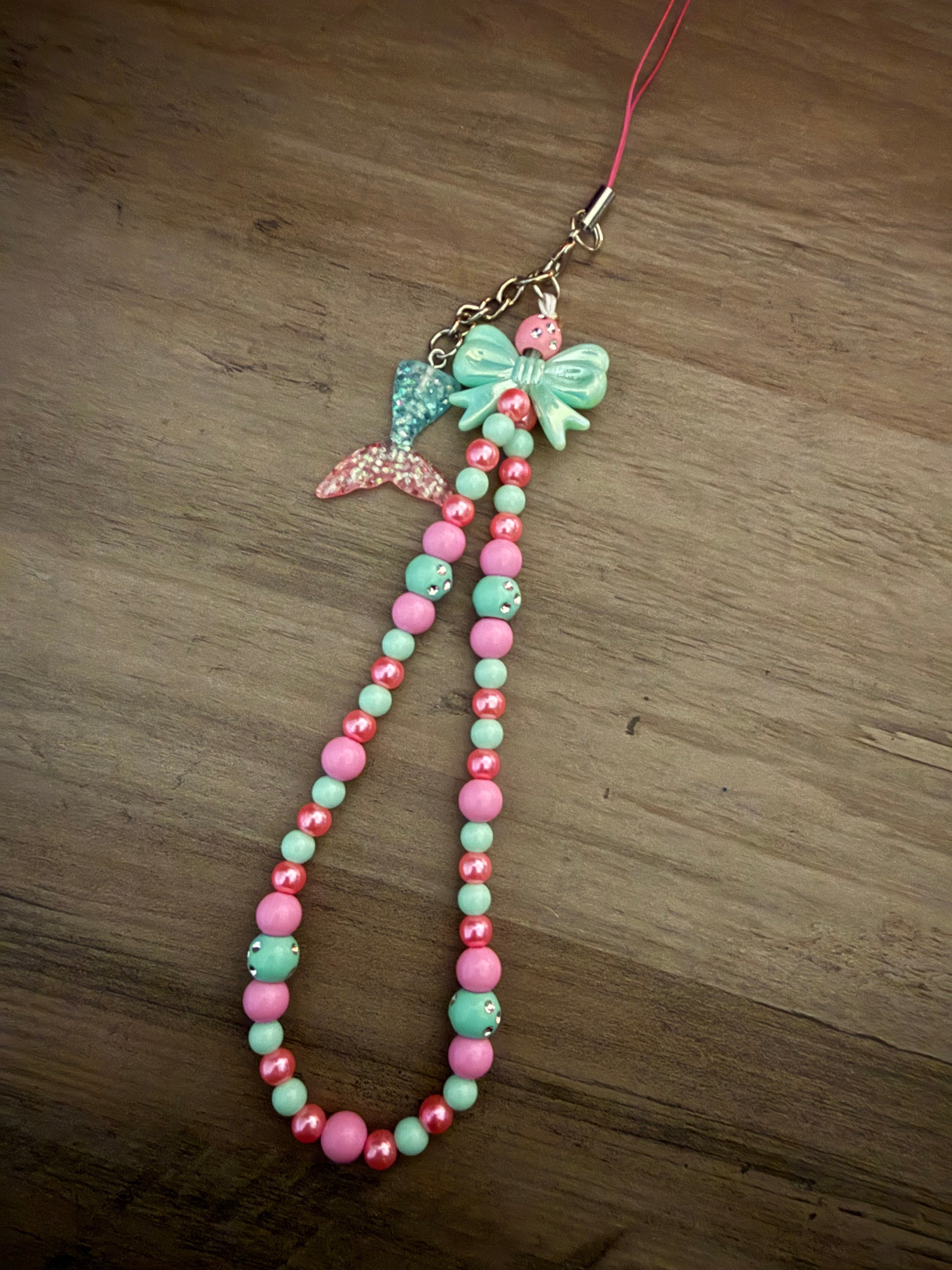 A beaded phone wristlet strap in shades of pink and mint green, featuring a matching green bow and a sparkling mermaid tail charm.