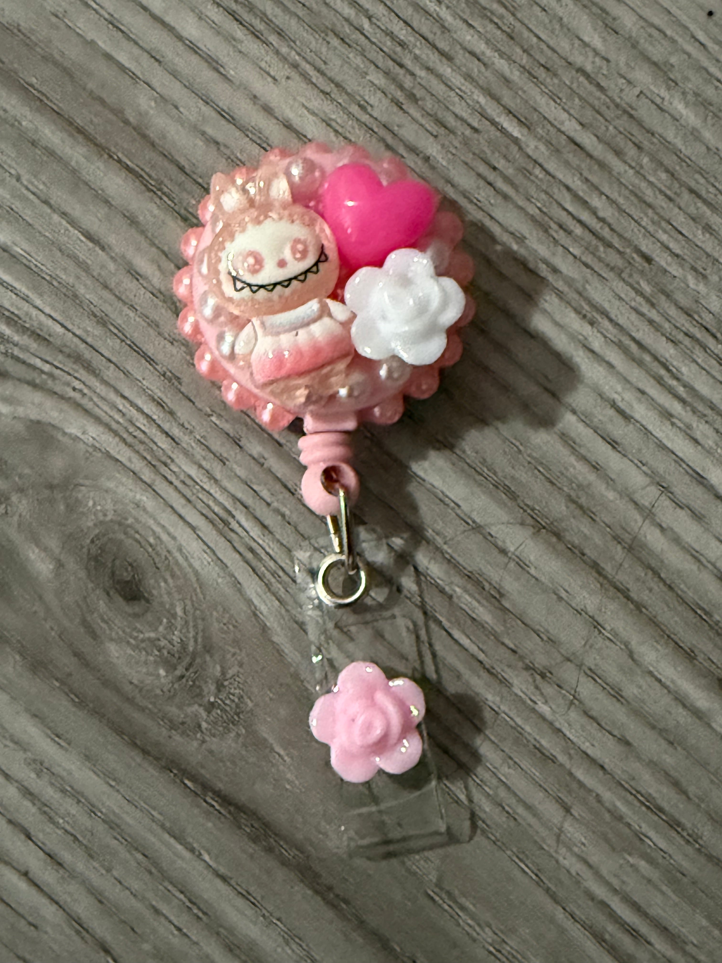 Cute pink badge reel featuring a whimsical monster with bunny ears, a pink heart, and white flowers, attached to a small pink flower charm.