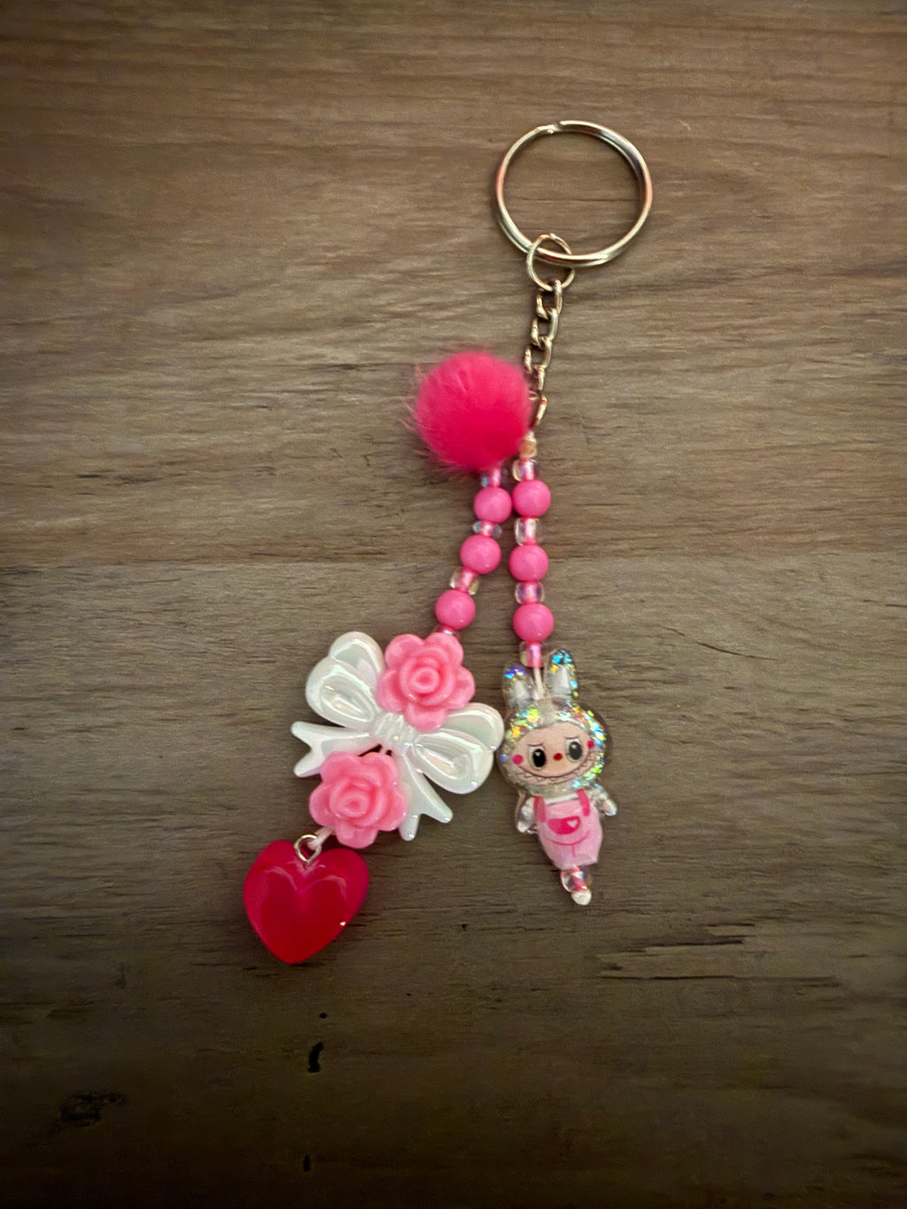 A pink beaded keychain featuring a fluffy pom-pom, a white bow with pink roses, a red heart charm, and a small glittery monster charm.