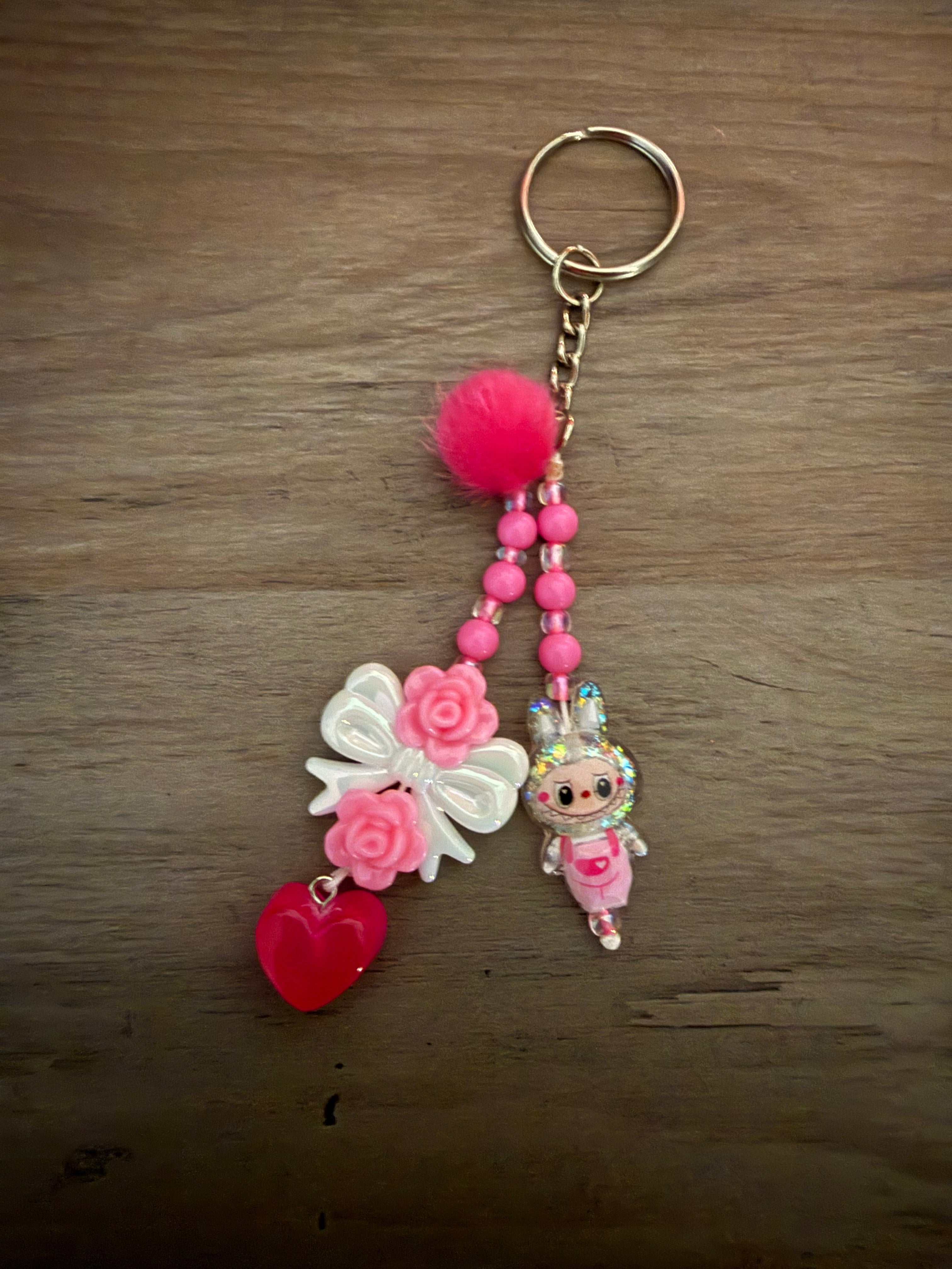 A pink beaded keychain featuring a fluffy pom-pom, a white bow with pink roses, a red heart charm, and a small glittery monster charm.
