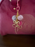 A handcrafted bag charm attached to a pink velvet purse. The charm features a gold-tone chain with three small pom-poms in grey, light pink, and hot pink, finished with two delicate gold-tone wire bows.