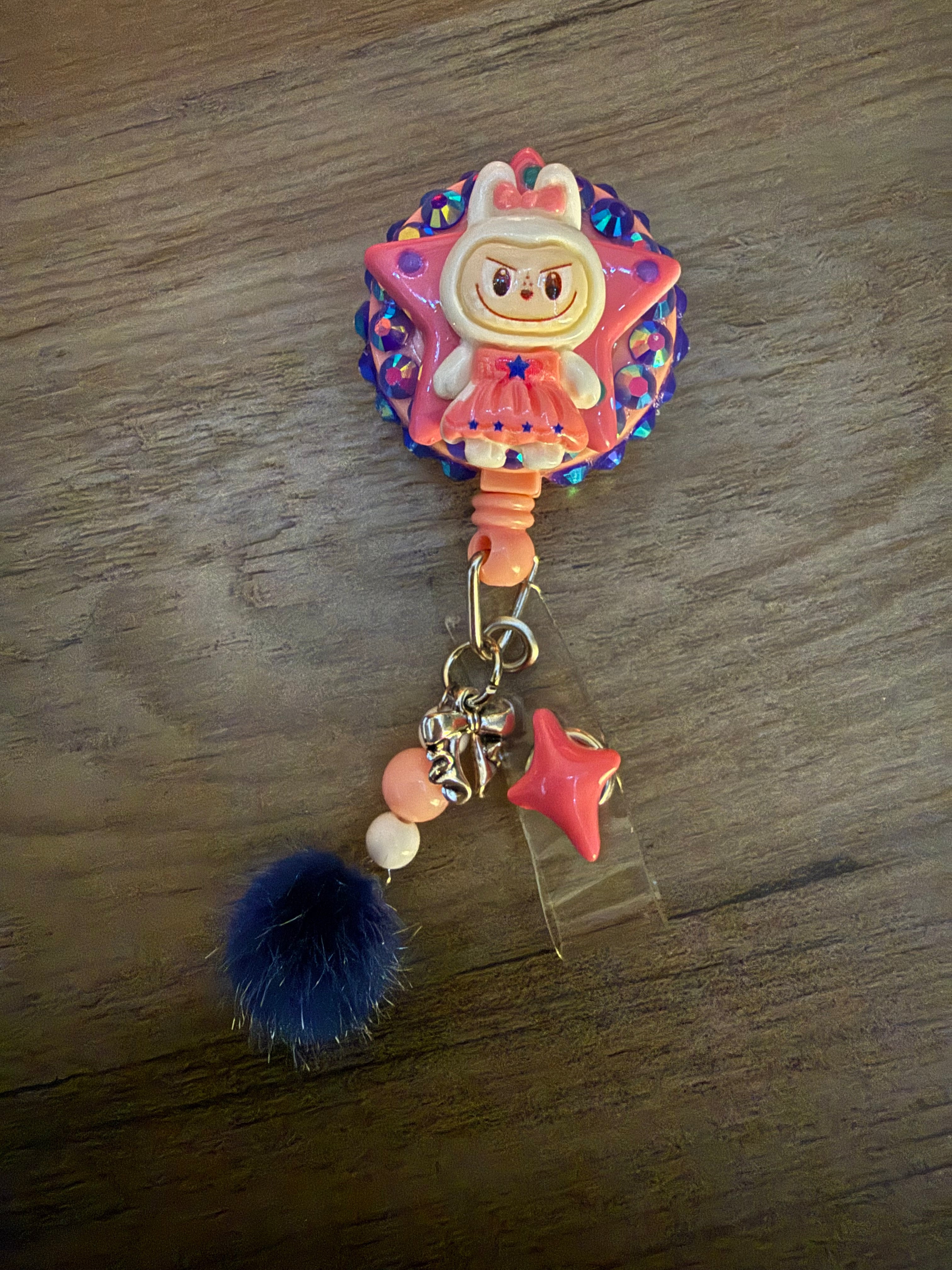 A playful badge reel featuring a cartoon monster character, star-emblazoned costume. The reel is adorned with rhinestones, a navy blue pom-pom, a small dark pink star charm, and a decorative key and lock, all attached with a clear plastic strap.