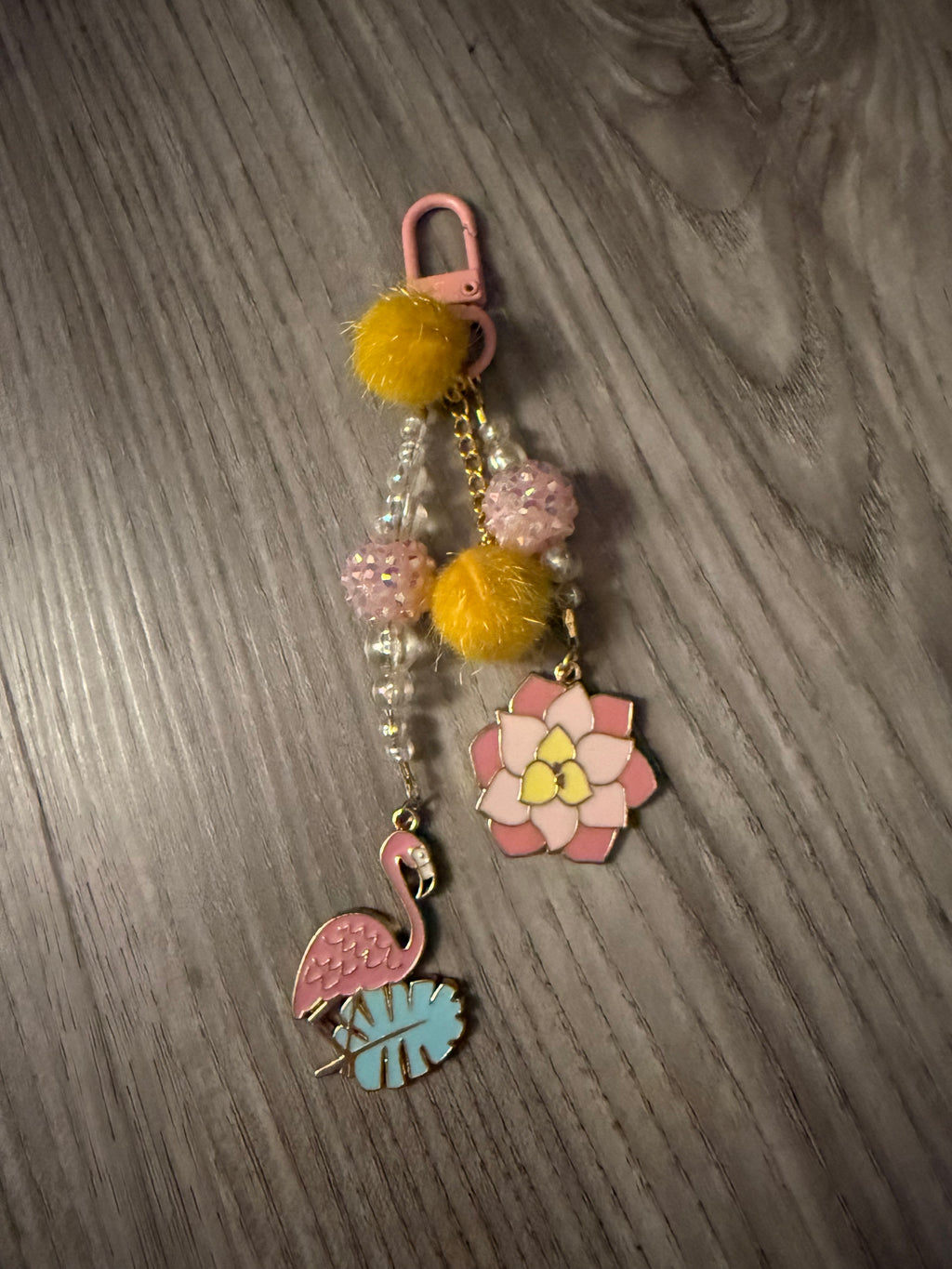 A handcrafted tropical-themed bag charm with a pink lobster clasp on a wood-grain background. The charm features a pink flamingo on a blue leaf, a pink and yellow lotus flower, yellow pom-poms, and a strand of iridescent and pink textured beads.