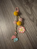 A handcrafted tropical-themed bag charm with a pink lobster clasp on a wood-grain background. The charm features a pink flamingo on a blue leaf, a pink and yellow lotus flower, yellow pom-poms, and a strand of iridescent and pink textured beads.