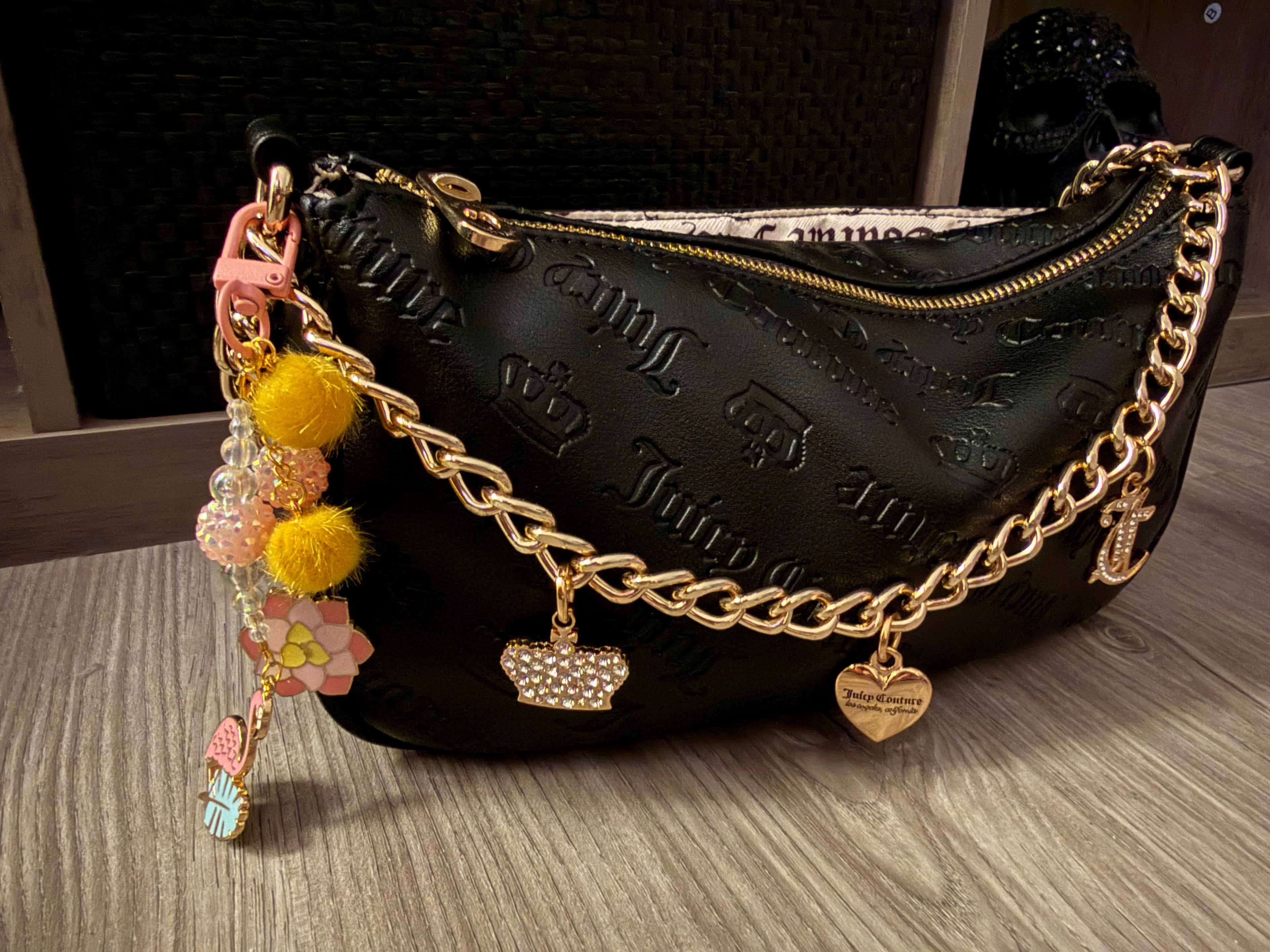 A black embossed Juicy Couture handbag with a gold-tone chain strap featuring crown and heart charms. A handcrafted tropical-themed bag charm with a pink flamingo, lotus flower, yellow pom-poms, and iridescent beads is attached to the side.