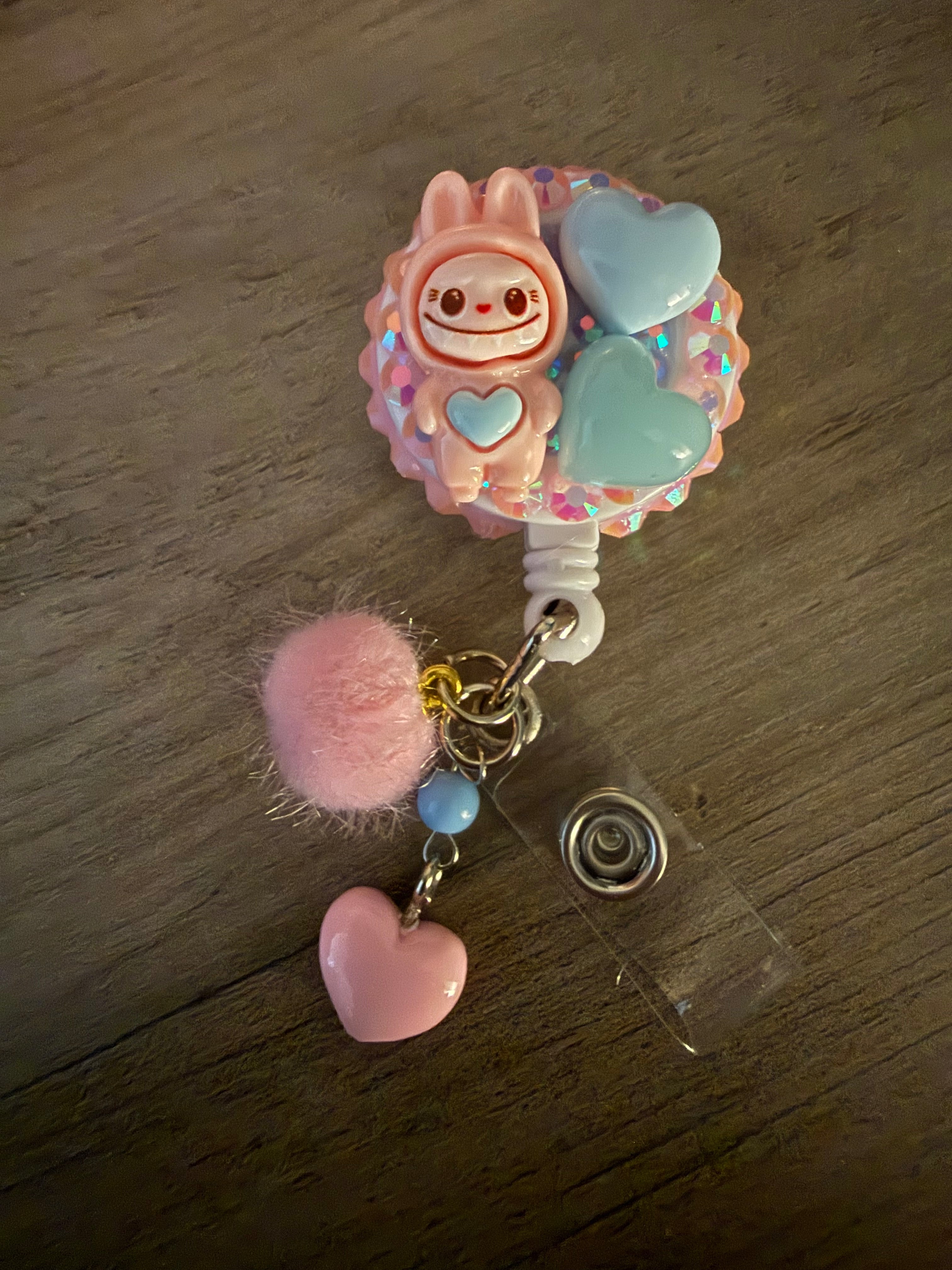 A close-up of a handcrafted pink retractable badge reel decorated with a monster in a bunny suit, blue heart accents, and rhinestones, featuring a pink pom-pom and heart charm.