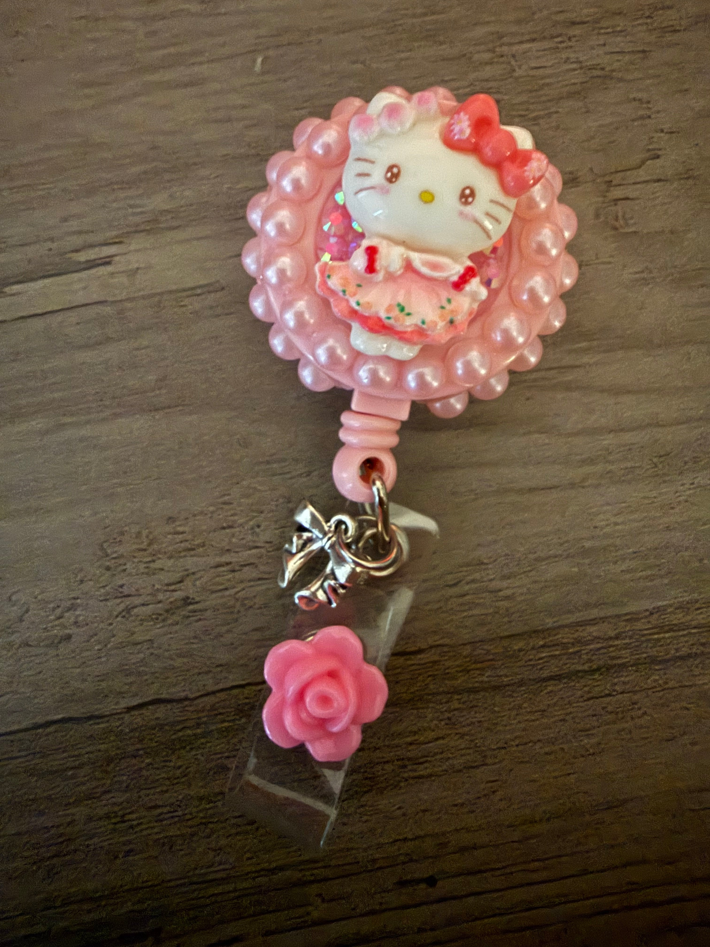 Colorful badge reel featuring a Hello Kitty inspired charm in a pink dress, surrounded by a pink beaded frame, with a small pink rose accent and a silver charm.