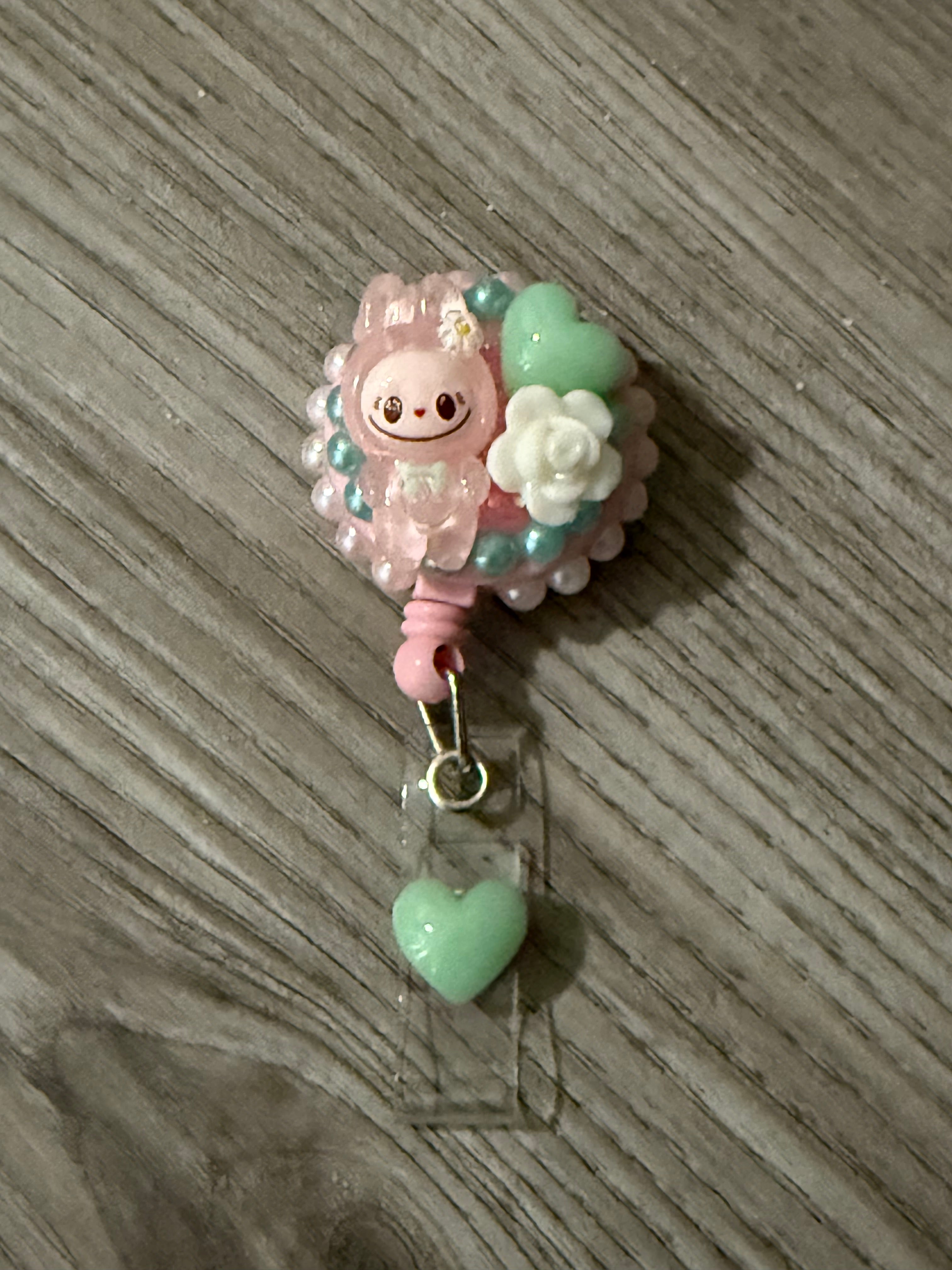 Cute pink badge reel featuring a whimsical character design, surrounded by pastel beads and floral accents, with a small green heart charm attached.