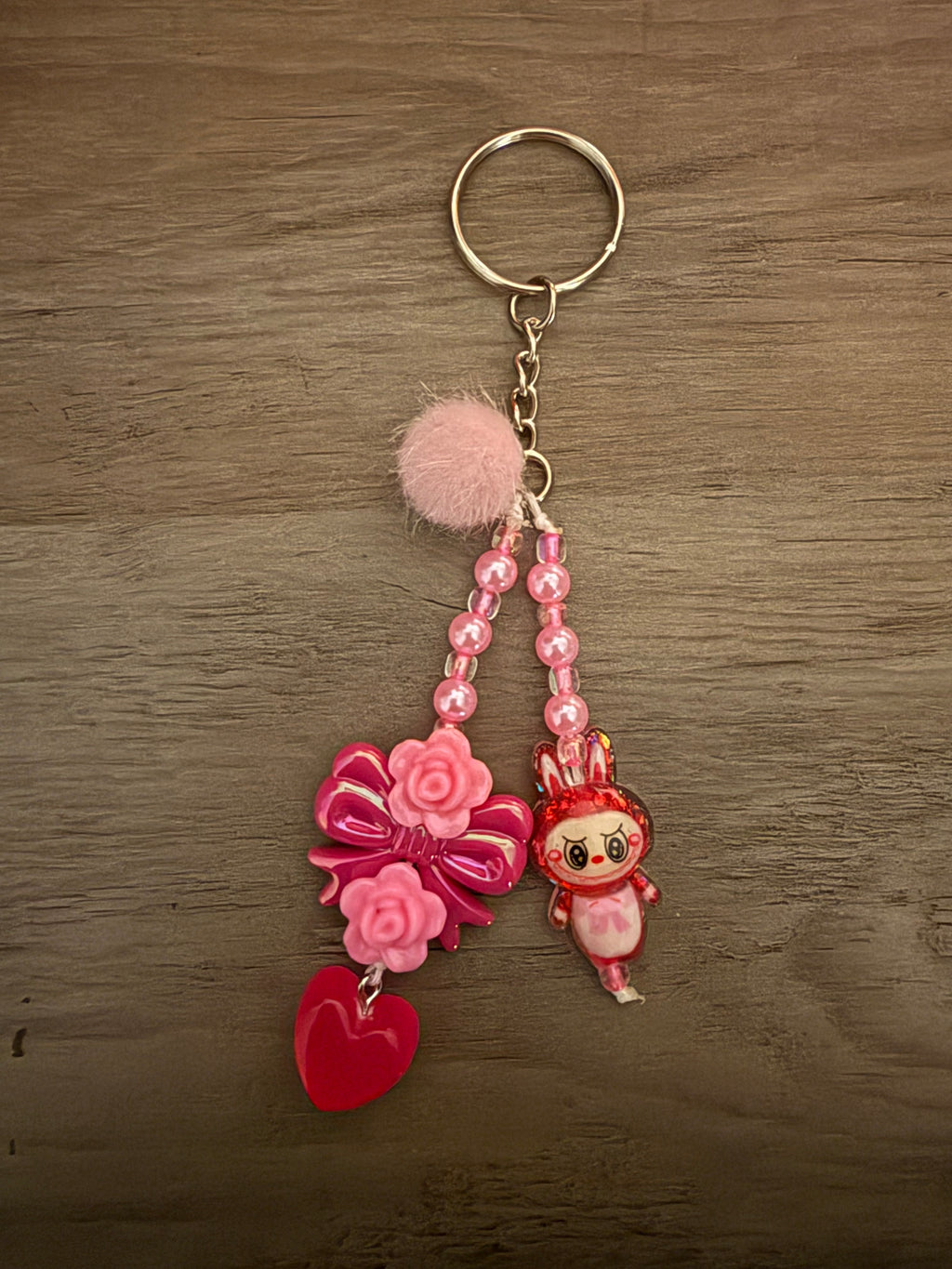 A handcrafted pink keychain featuring two beaded strands hanging from a silver ring. The strands include a pink pom-pom, a cartoon bunny monster, a large pink bow decorated with roses, and a translucent red heart charm.