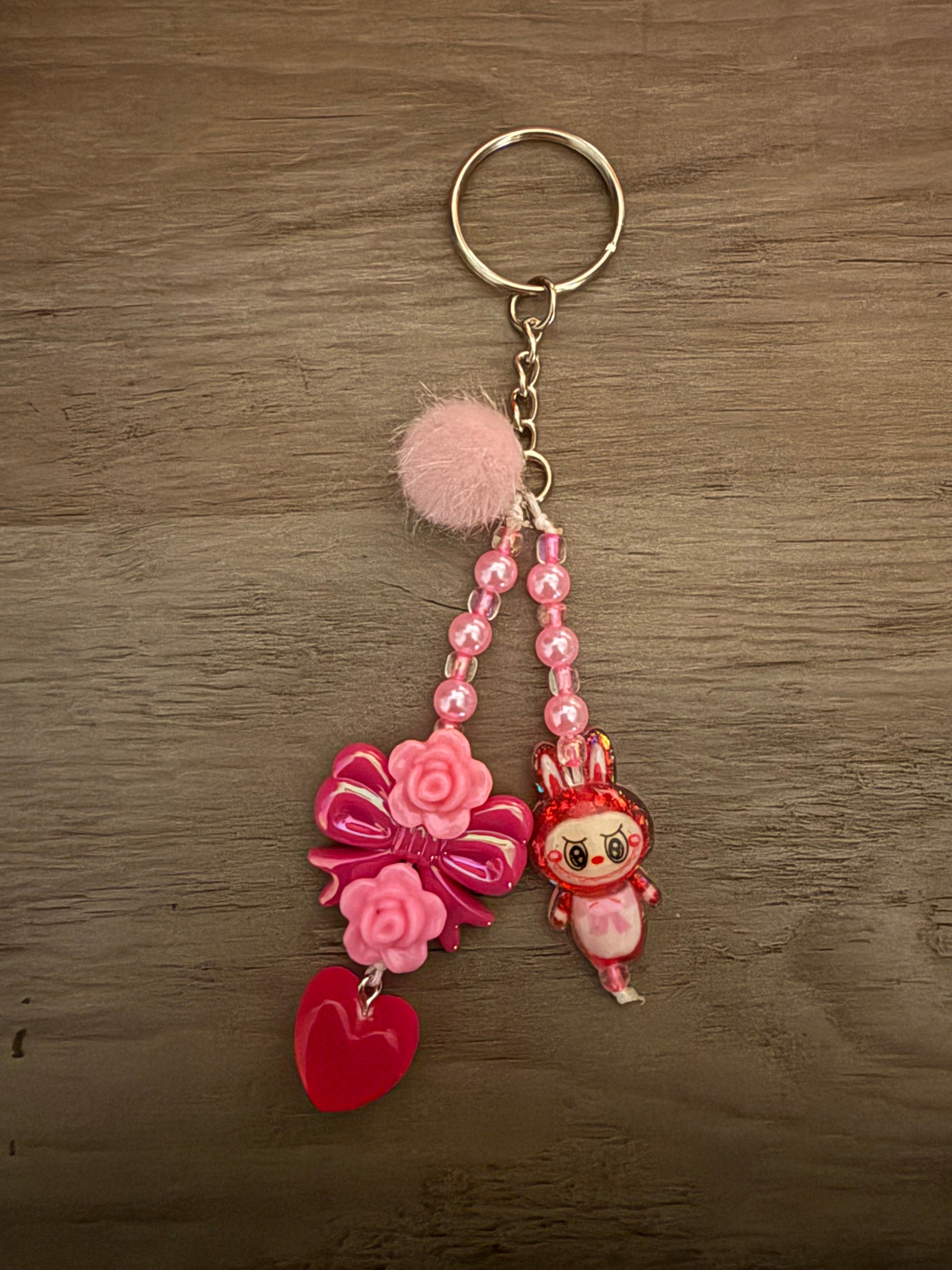 A handcrafted pink keychain featuring two beaded strands hanging from a silver ring. The strands include a pink pom-pom, a cartoon bunny monster, a large pink bow decorated with roses, and a translucent red heart charm.