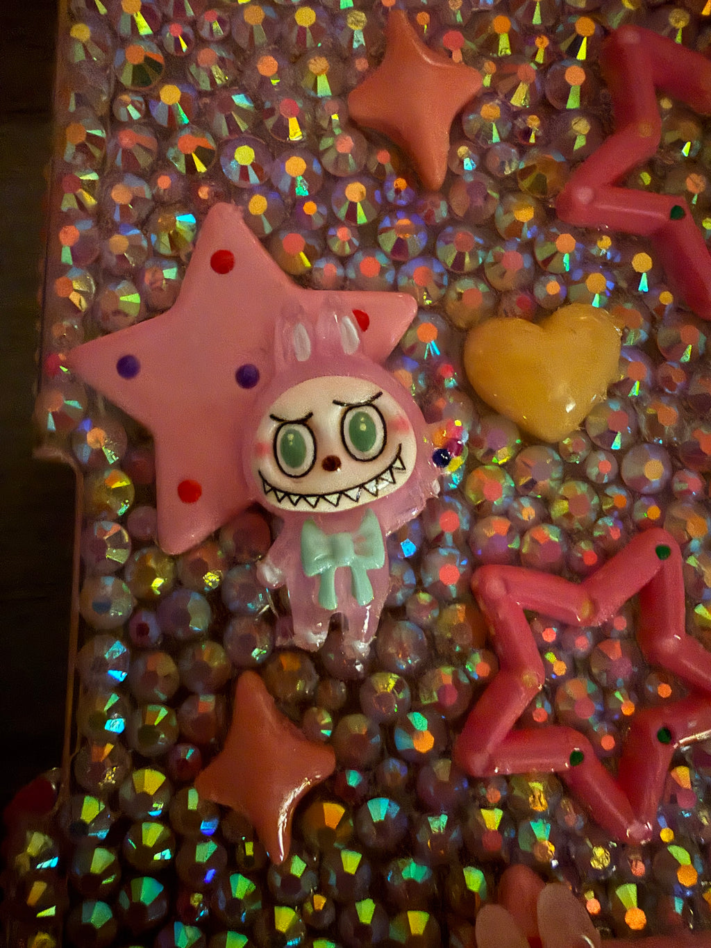 Close-up of a decoden-style surface covered in iridescent rhinestones, featuring a pink star and a cartoonish pink monster charm with a wide, toothy grin and a mint green bow.
