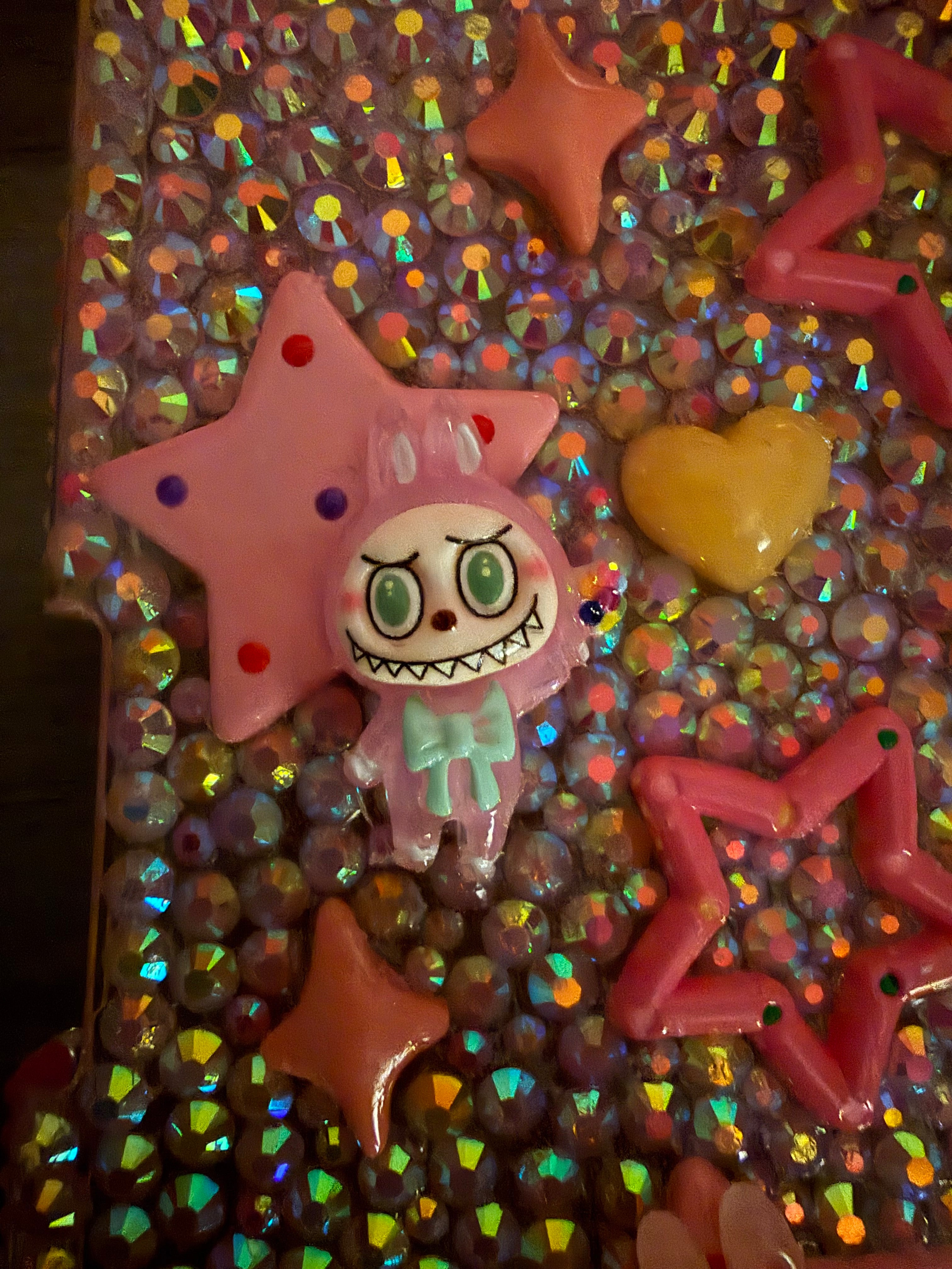 Close-up of a decoden-style surface covered in iridescent rhinestones, featuring a pink star and a cartoonish pink monster charm with a wide, toothy grin and a mint green bow.