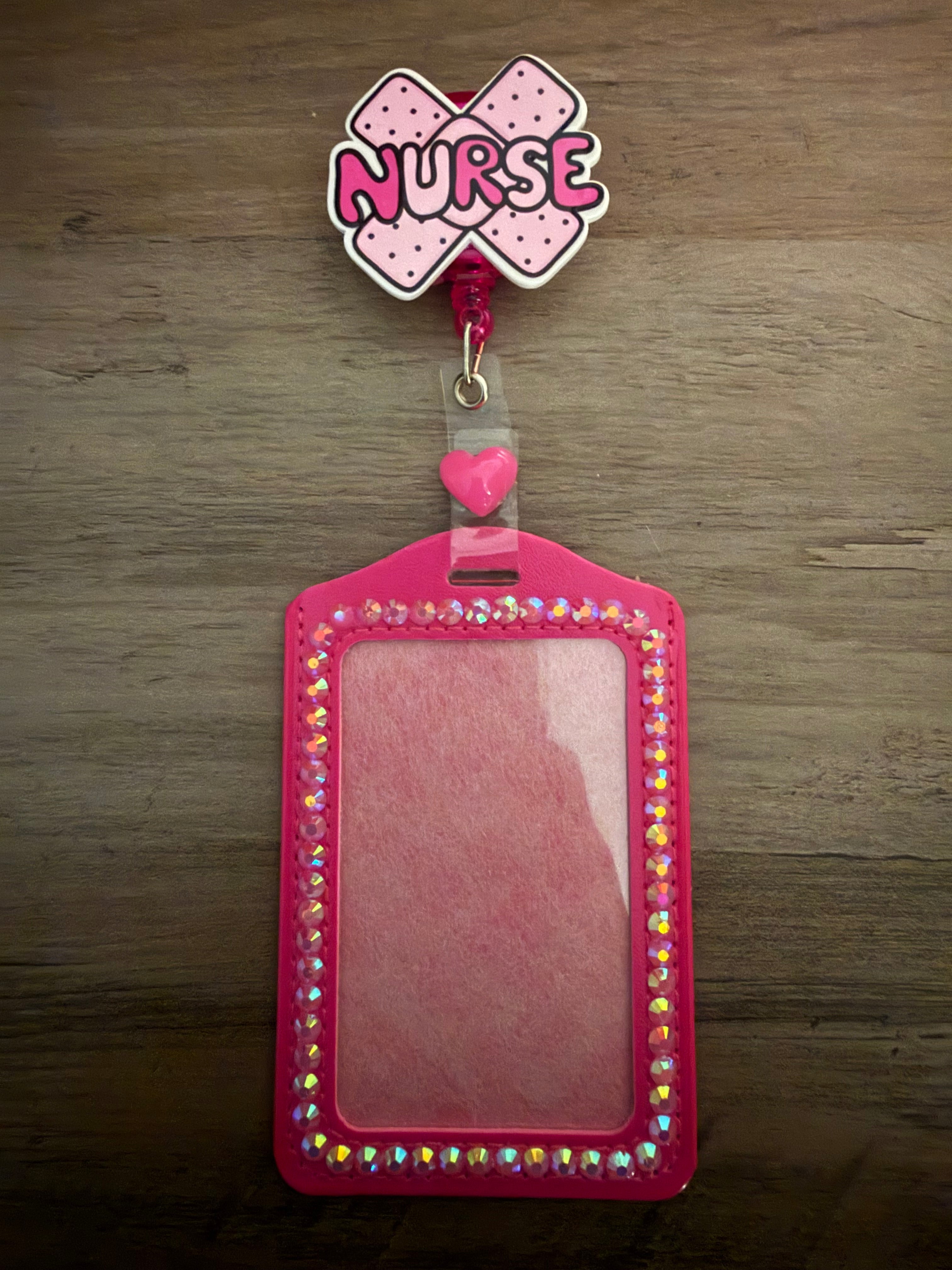 A bright pink, rhinestone-lined ID holder attached to a retractable badge reel. The reel is shaped like two crossed pink bandages with the word "NURSE" written across them, accented by a small pink heart charm.