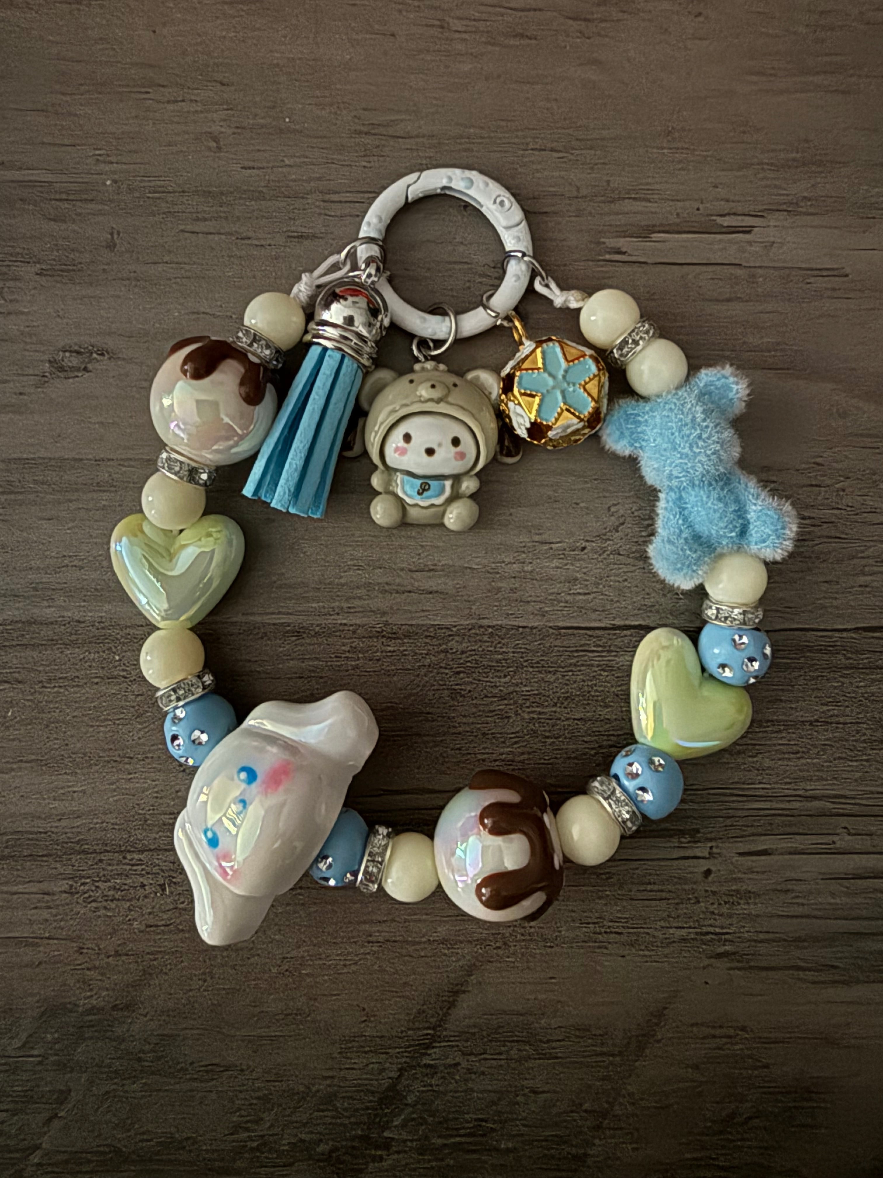 A pastel blue and cream beaded charm featuring Pochacco and Cinnamoroll figures, a fuzzy blue bear, and a blue tassel. The beads include cream rounds, pale green hearts, chocolate-drizzled accents, and rhinestone-studded blue spacers, all attached to a white circular clasp.
