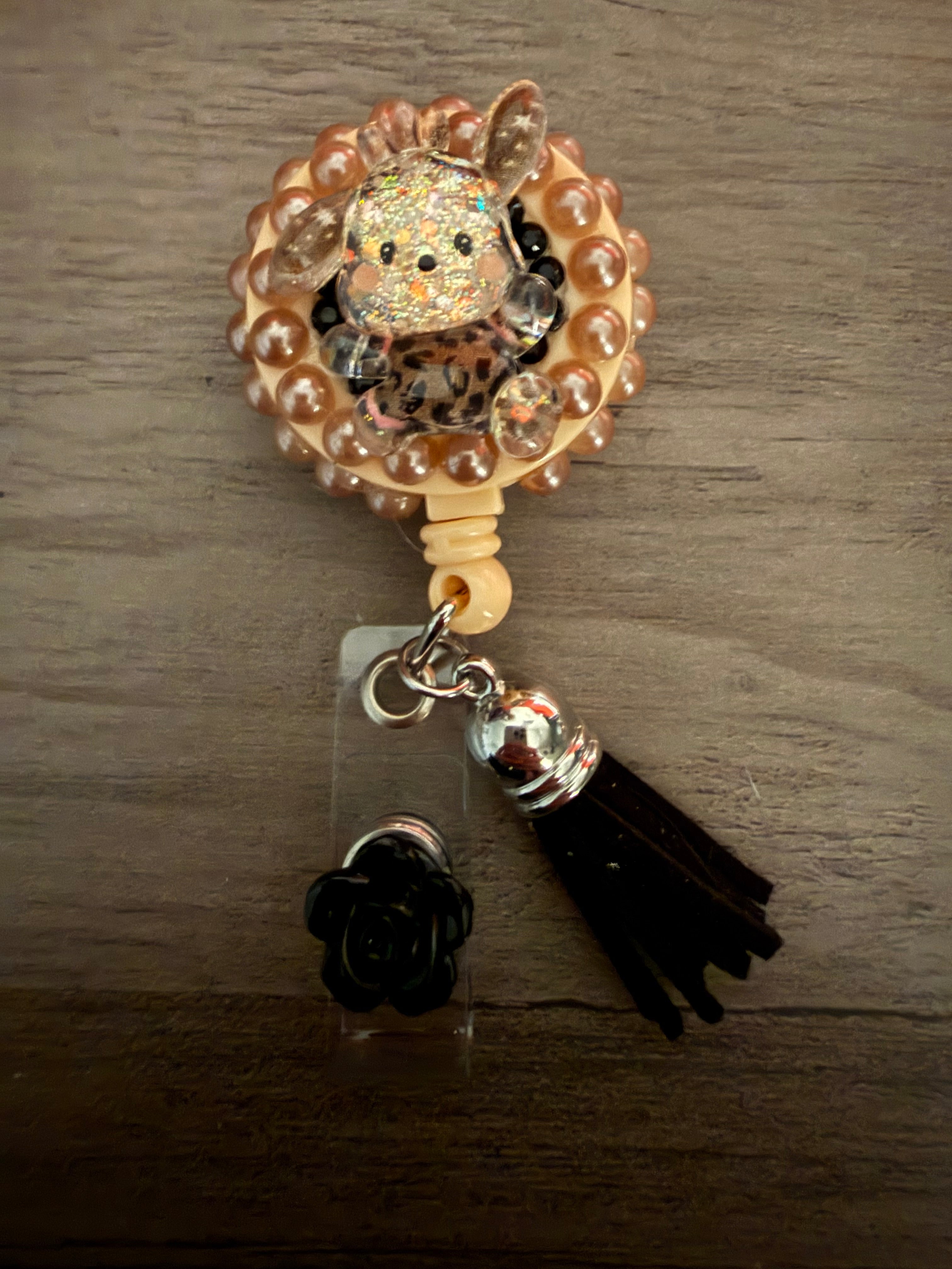 A decoden retractable badge reel featuring a glittery Pochacco character in leopard print, bordered by bronze pearls and finished with a black tassel and black rose charm.