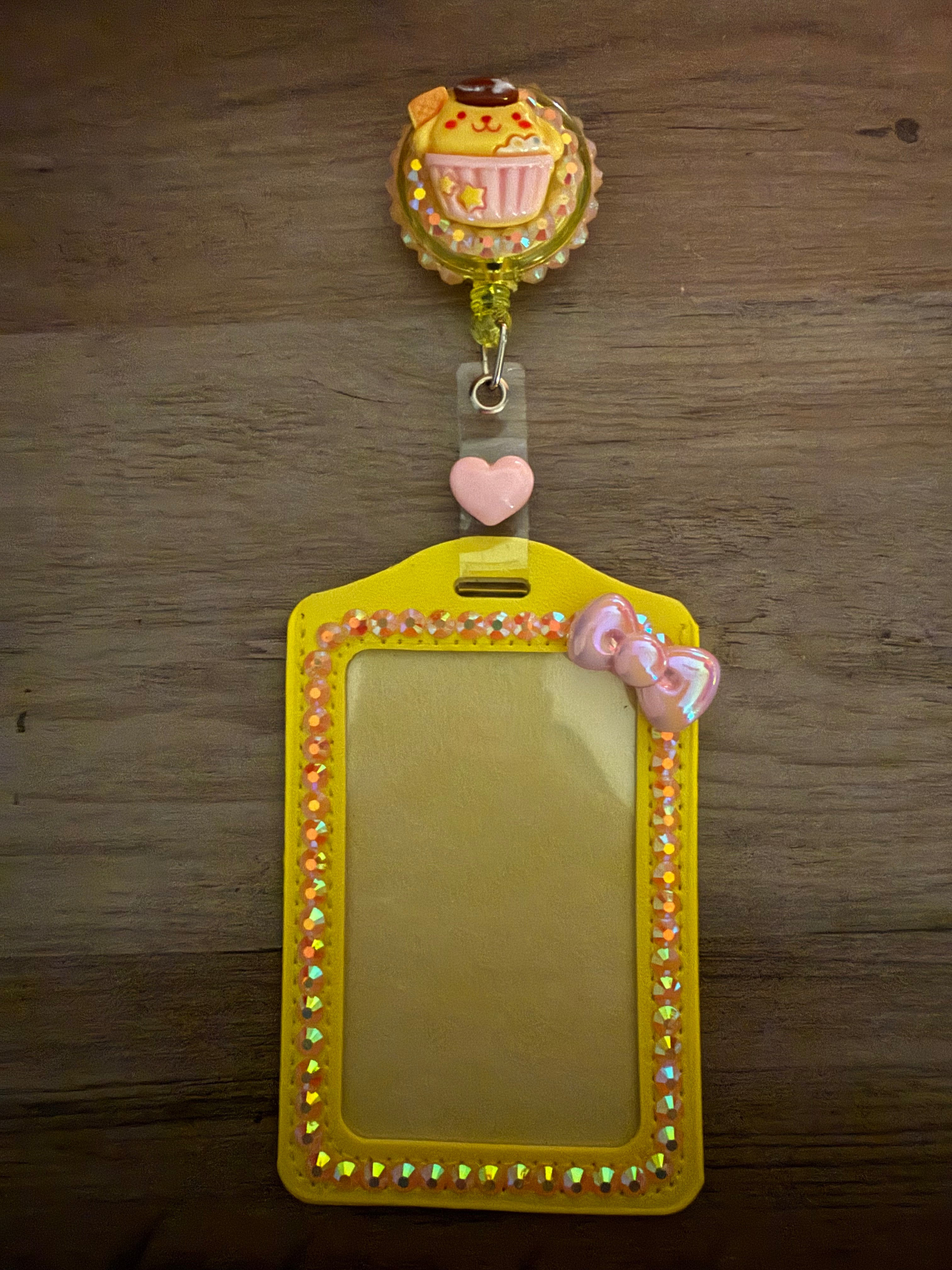 A yellow, rhinestone-embellished ID card holder with a matching retractable badge reel. The reel features a Pompompurin cupcake charm set against a shimmering background, and the holder is accented with iridescent gems and a small pink pearlescent bow.