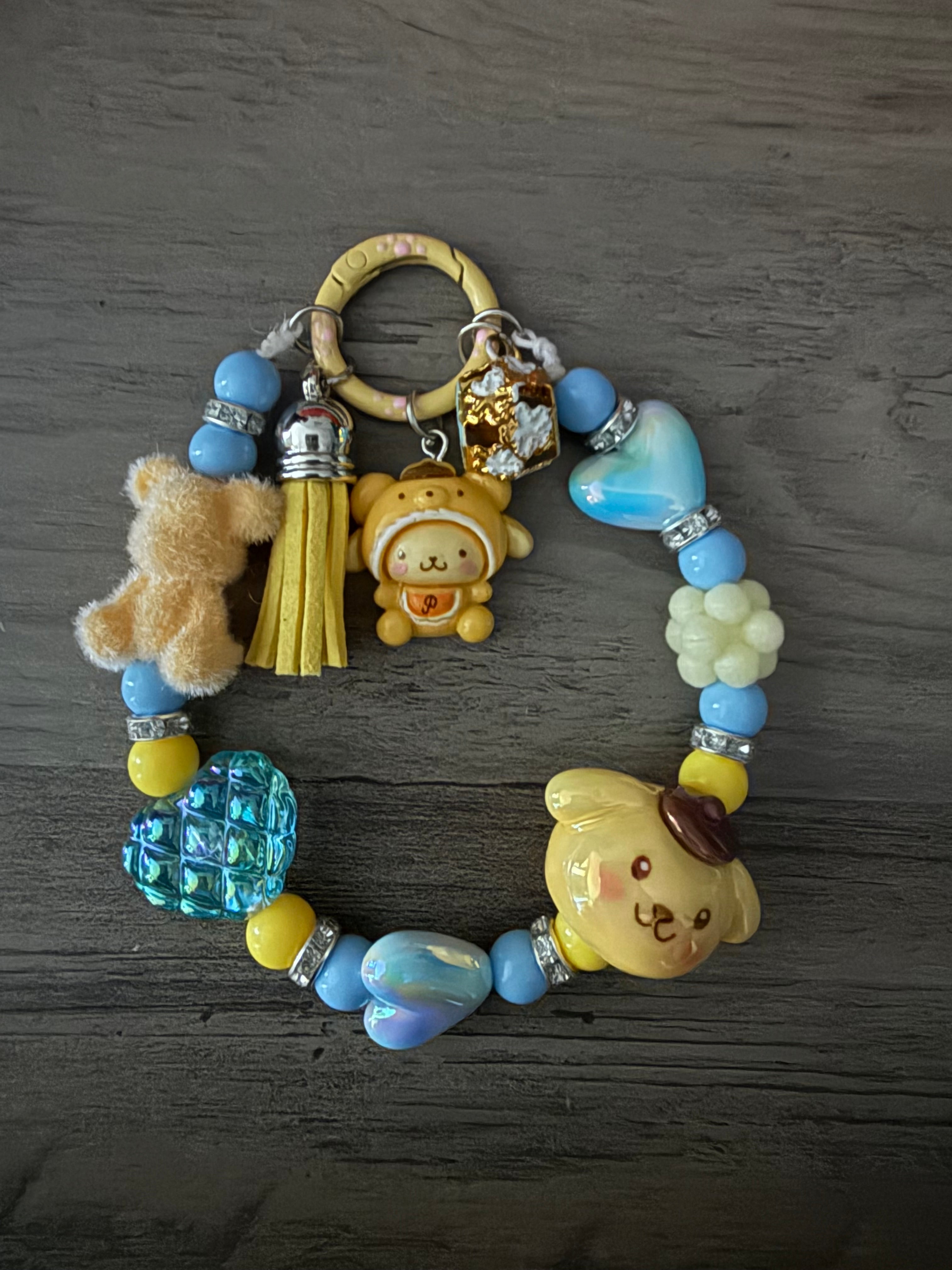 A pastel-themed beaded charm featuring various Pompompurin figures, a fuzzy tan bear, and a yellow tassel. The beads include round blue and yellow accents, iridescent blue hearts, and silver rhinestone spacers, all attached to a pale yellow circular clasp.
