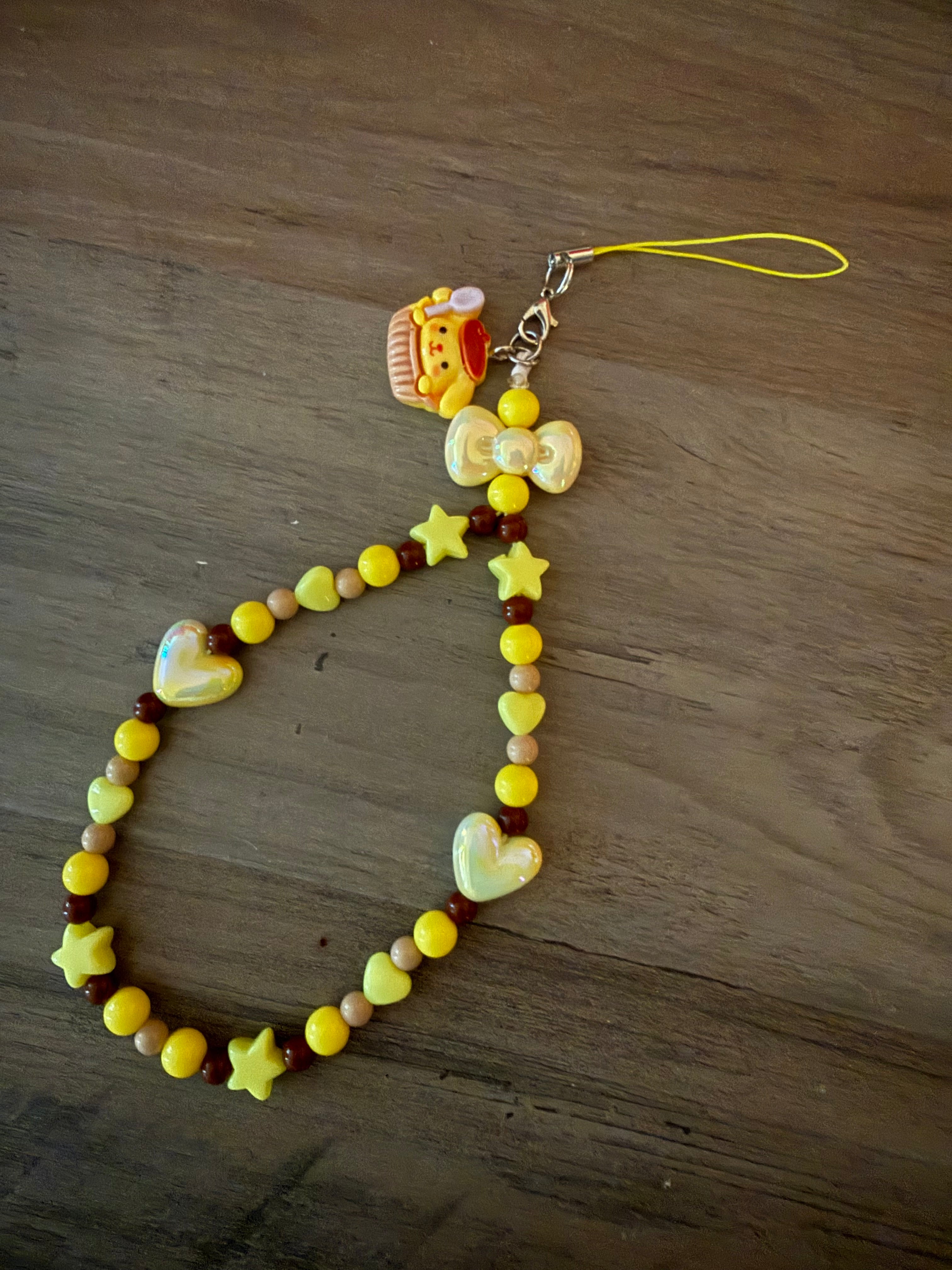 A handcrafted beaded phone wristlet featuring a mix of yellow, brown, and tan beads with yellow star and heart charms, finished with a yellow bow and a Pompompurin cupcake charm.