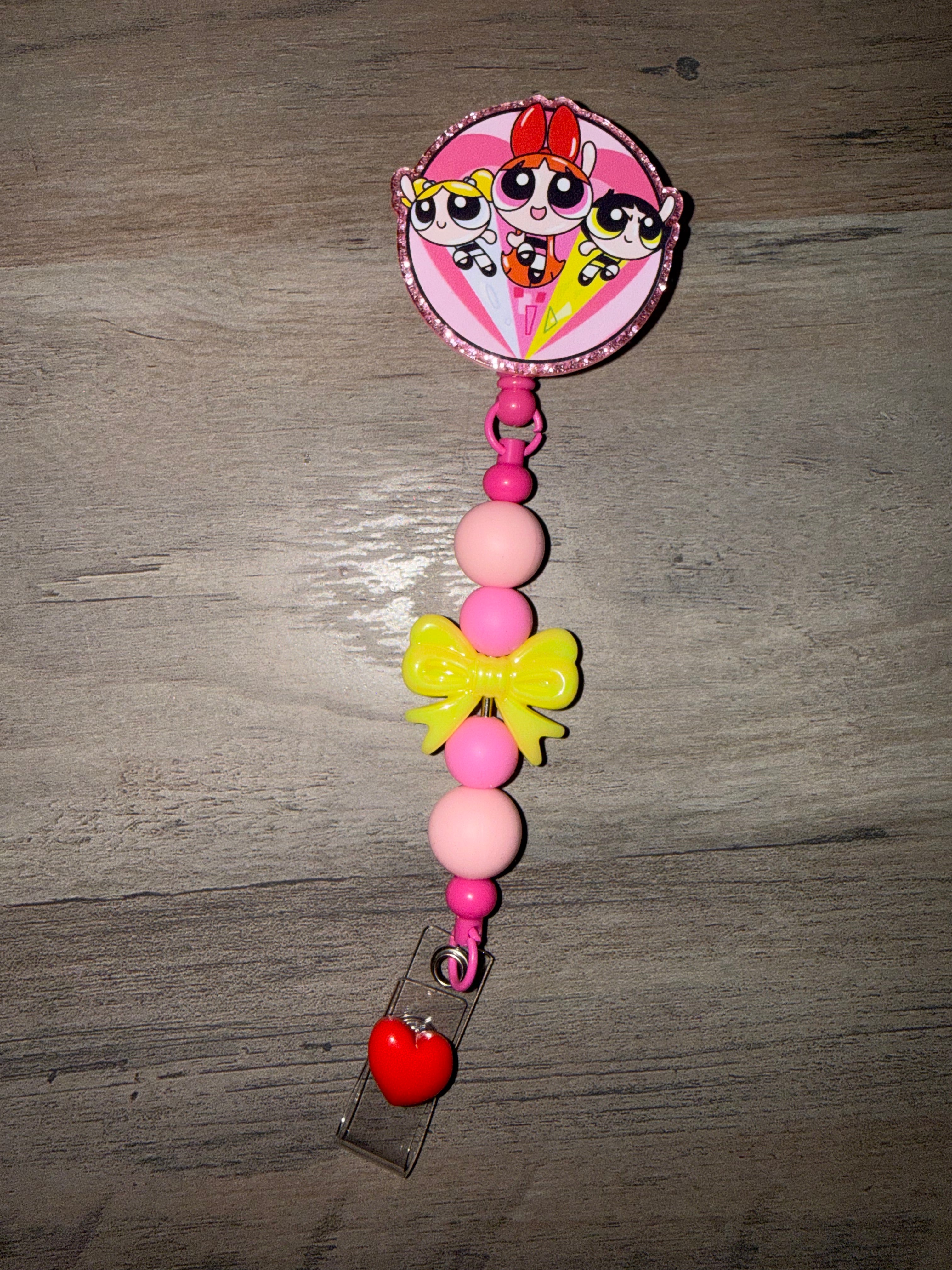 A handcrafted retractable badge reel featuring a glittery Powerpuff Girls charm with Blossom, Bubbles, and Buttercup. The reel is decorated with light and dark pink beads, a bright yellow bow, and a red heart accent at the bottom clip.