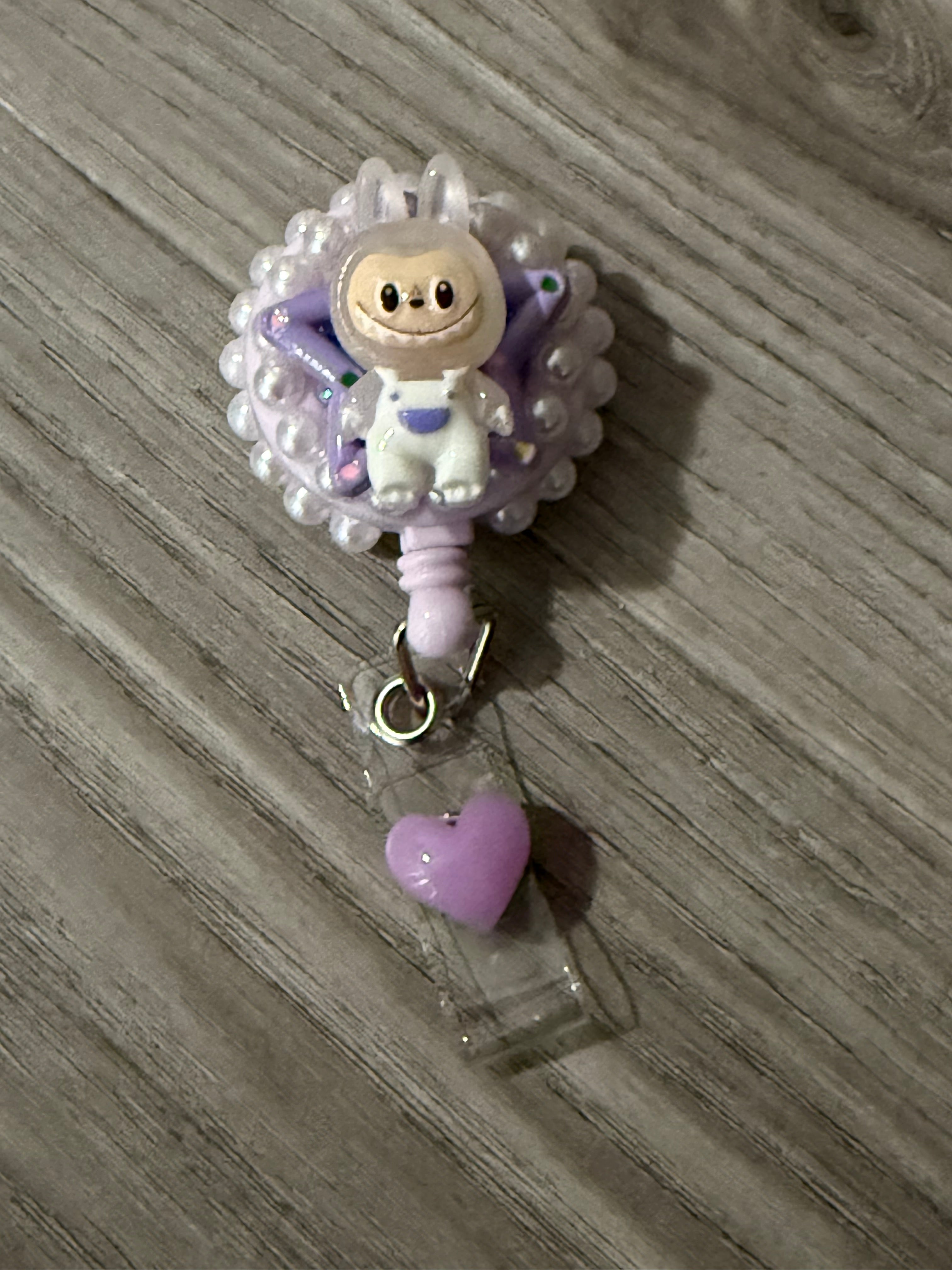 Cute badge reel featuring a whimsical monster in a purple bunny suit with a cheerful expression, surrounded by a decorative purple backing and a small pink heart charm.